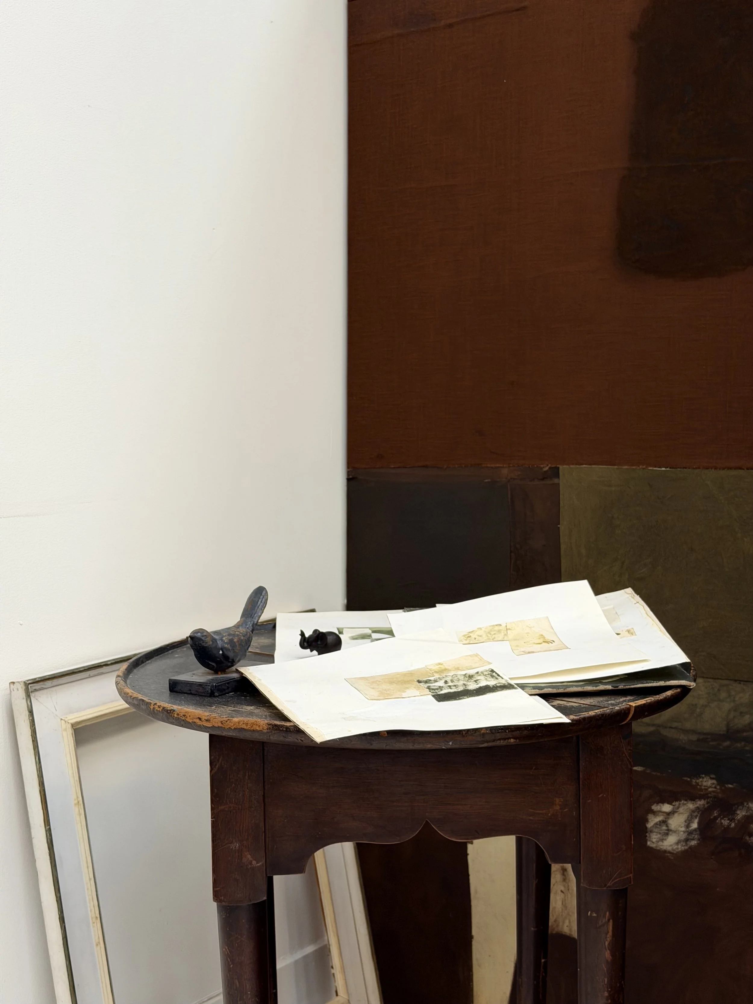 A small, round wooden table with several papers spread out on top. There is a black bird figurine and a black vase on the table. Behind the table is a large, dark wooden cabinet or panel and some picture frames leaning against the wall.