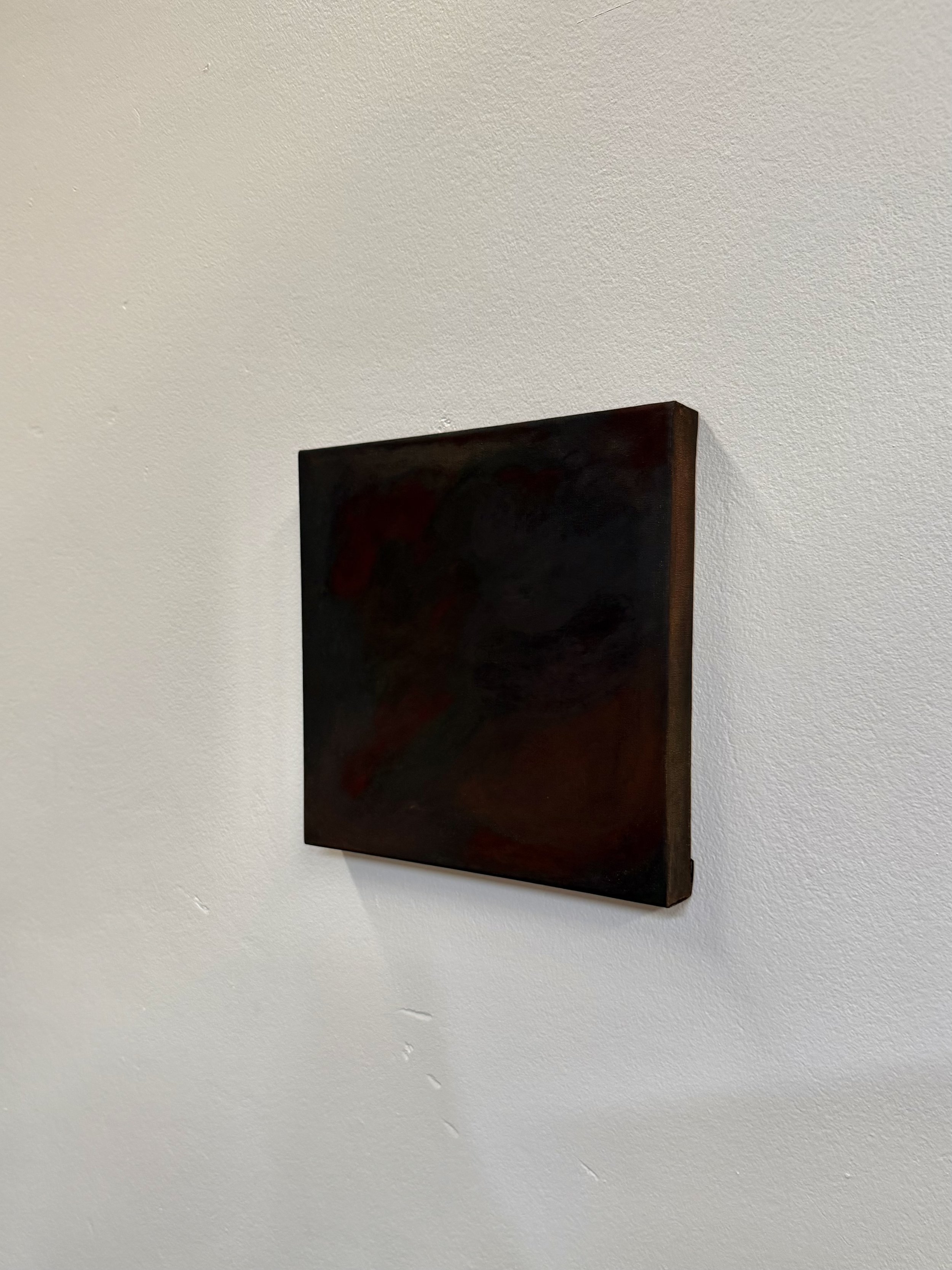 Small dark-colored painting with red and black abstract shapes, mounted on a white textured wall.