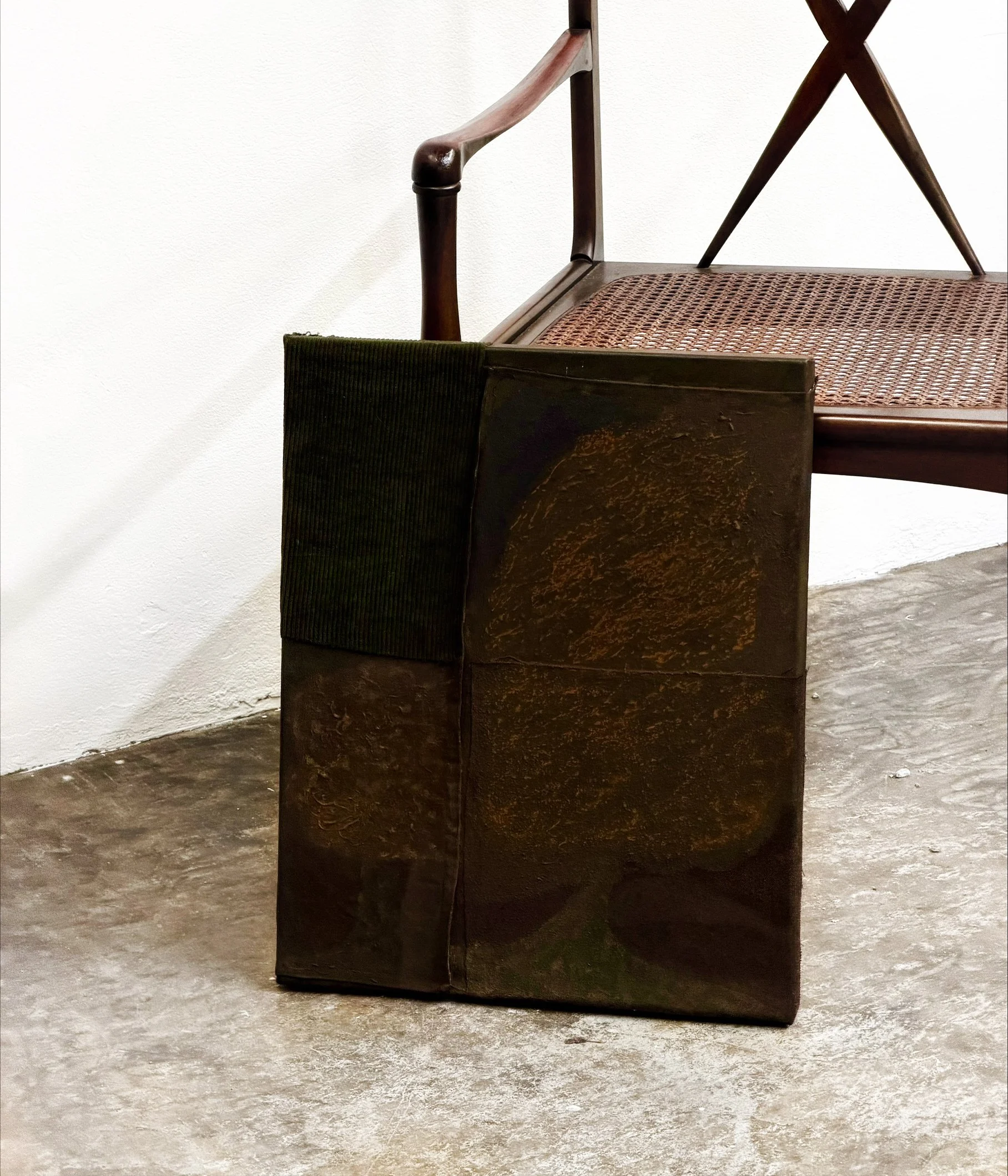 A wooden chair with a cane seat and curved armrests, positioned against a plain white wall, on a concrete floor.