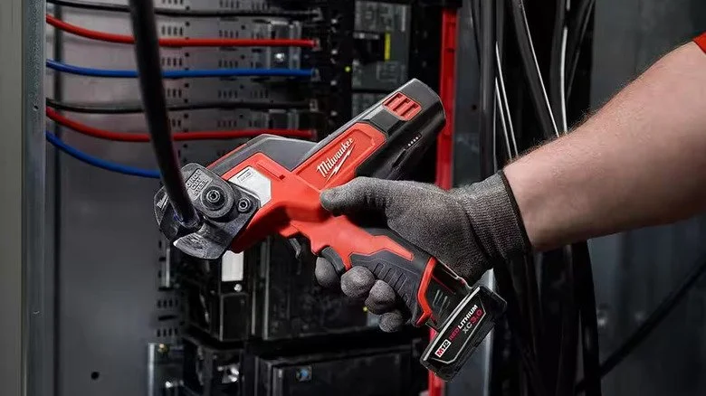 Person wearing a gray glove holding a Milwaukee cordless electrical tool near a power panel with red and blue wires.