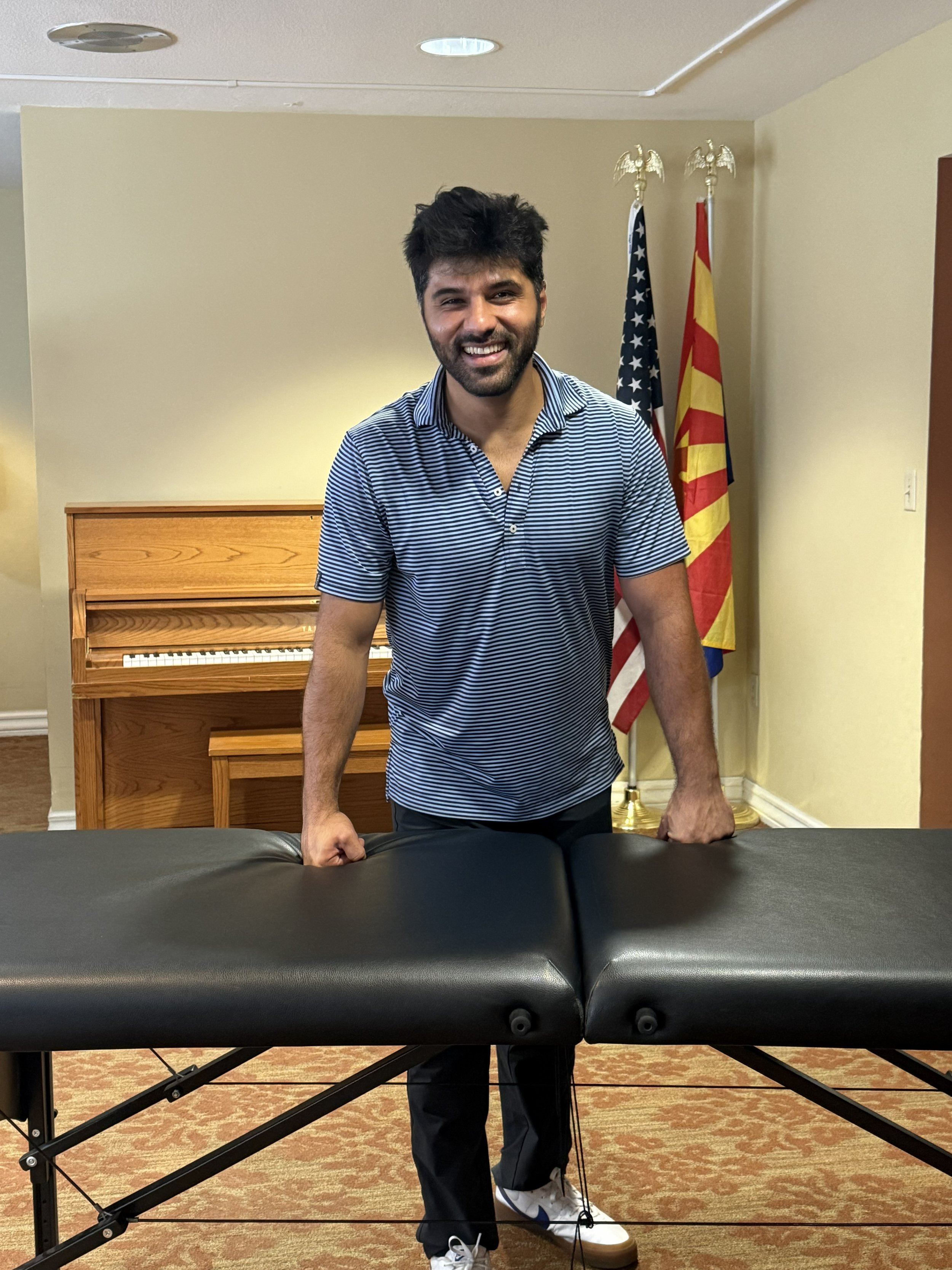 A smiling man in a blue and white striped shirt standing behind a massage table, with flags of the United States and another country in the background.