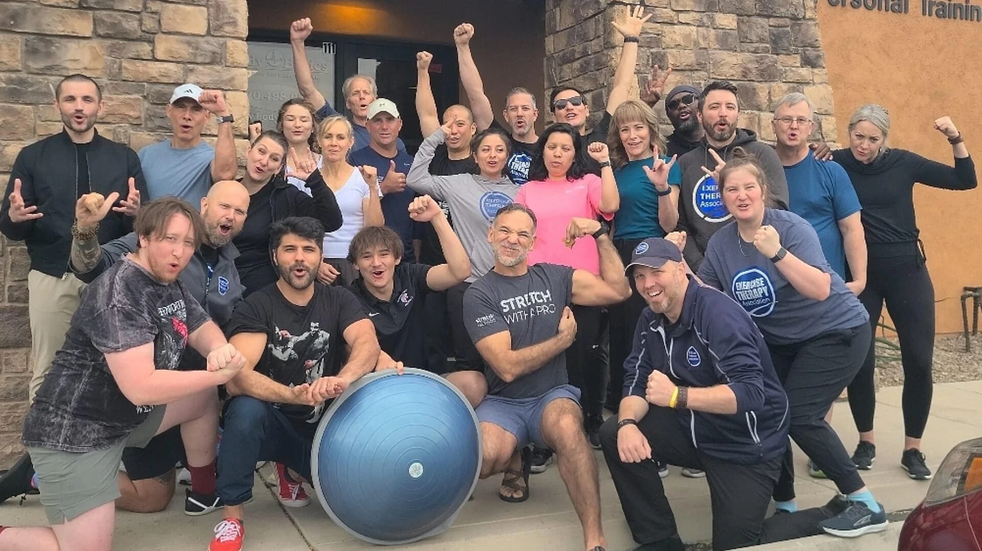 Group of diverse people celebrating outdoors, some flexing muscles, making fists, or cheering, in front of a stone and orange wall.