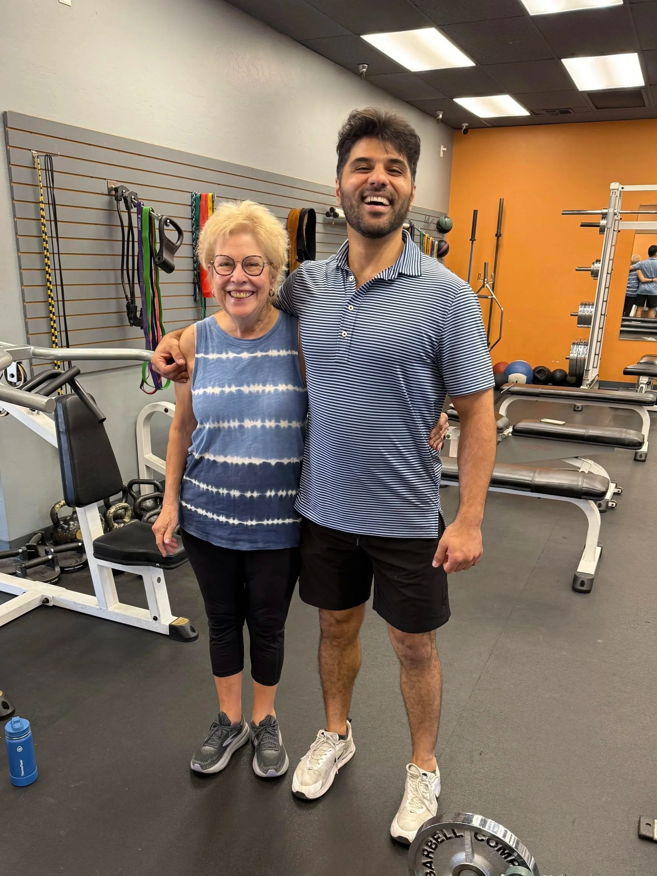 A middle-aged woman and a young man smiling at the camera in a gym, standing among various exercise equipment and weights.