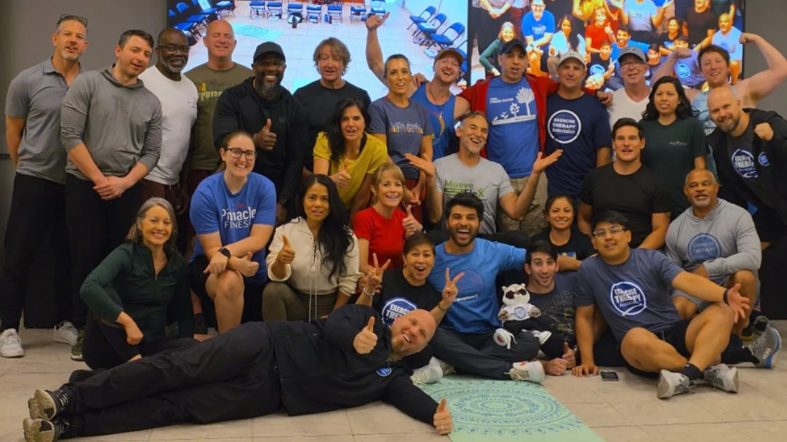 A large group of diverse people gathered indoors, smiling and posing for a group photo, some making peace signs or flexing muscles.