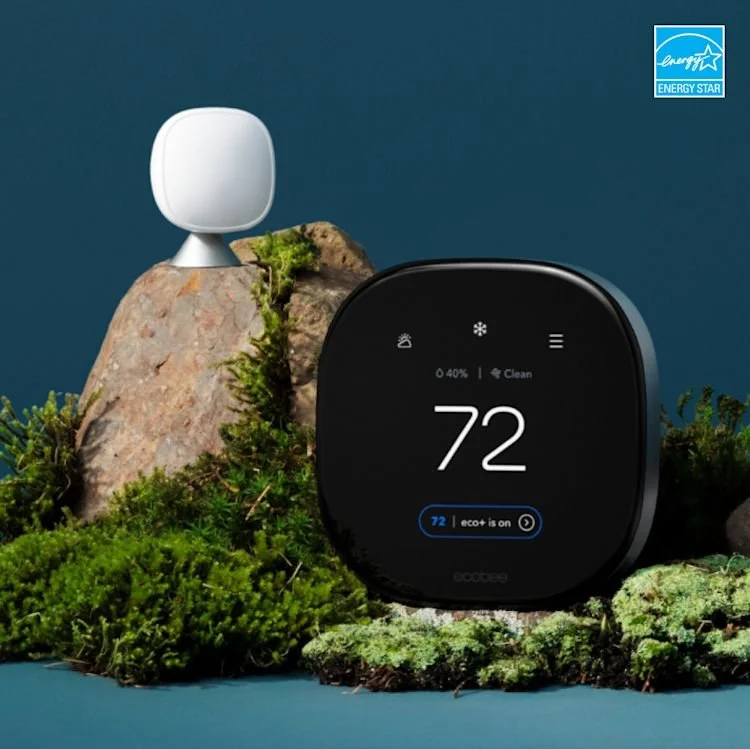 A Ecobee smart thermostat displaying a temperature of 72 degrees, surrounded by moss and rocks, with a room sensor on top of a rock and an Energy Star logo in the top right corner.