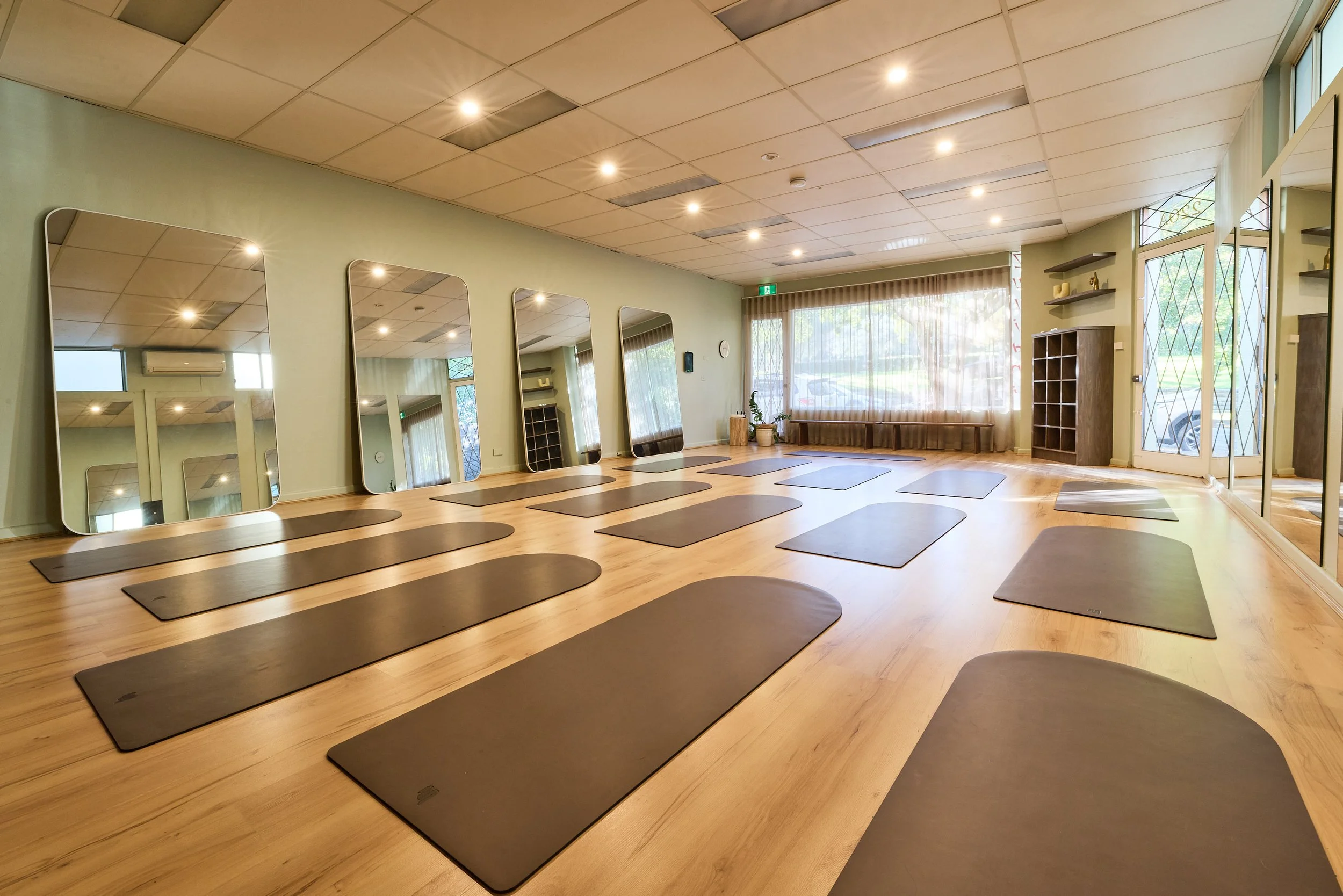 A spacious yoga studio with black mats arranged in rows on a wooden floor. The room has a large window with sheer curtains, multiple mirrors on one wall, and minimalist decor. The ceiling features recessed lighting.