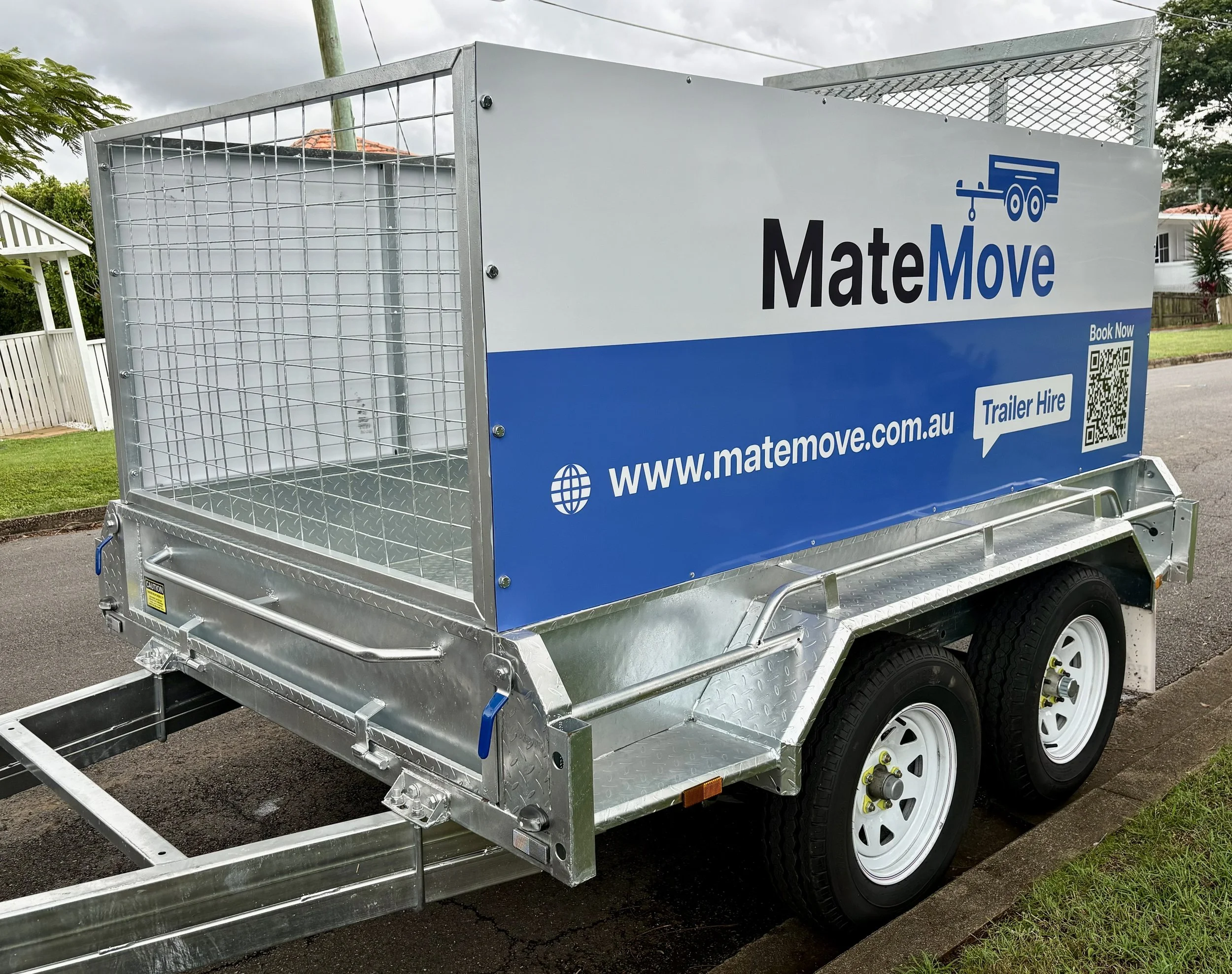 A metal trailer with a sign that reads 'MateMove' and includes a QR code, website link, and booking options, parked on a street with houses and trees in the background.