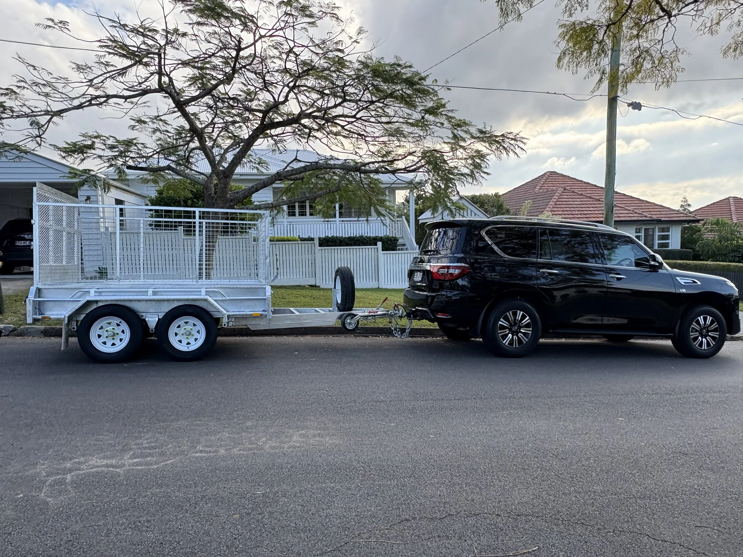 A black SUV is parked on the street behind a white trailer attached to a vehicle. There is a large tree with sparse leaves and a power pole with wires in the background. Houses with red and gray roofs are visible behind the trees.