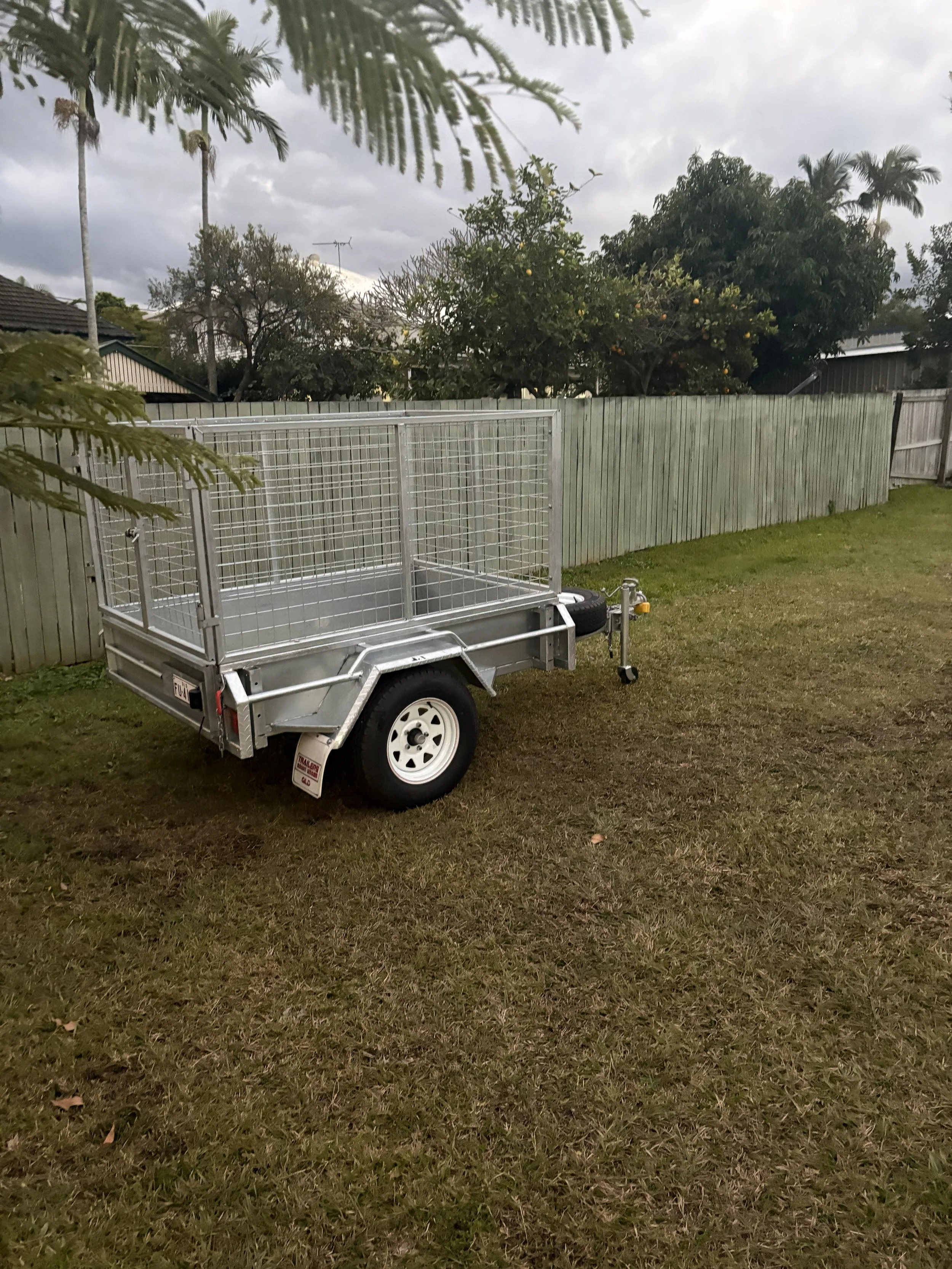 An outdoor scene with a metal trailer on grass, a wooden fence, trees, and cloudy sky.