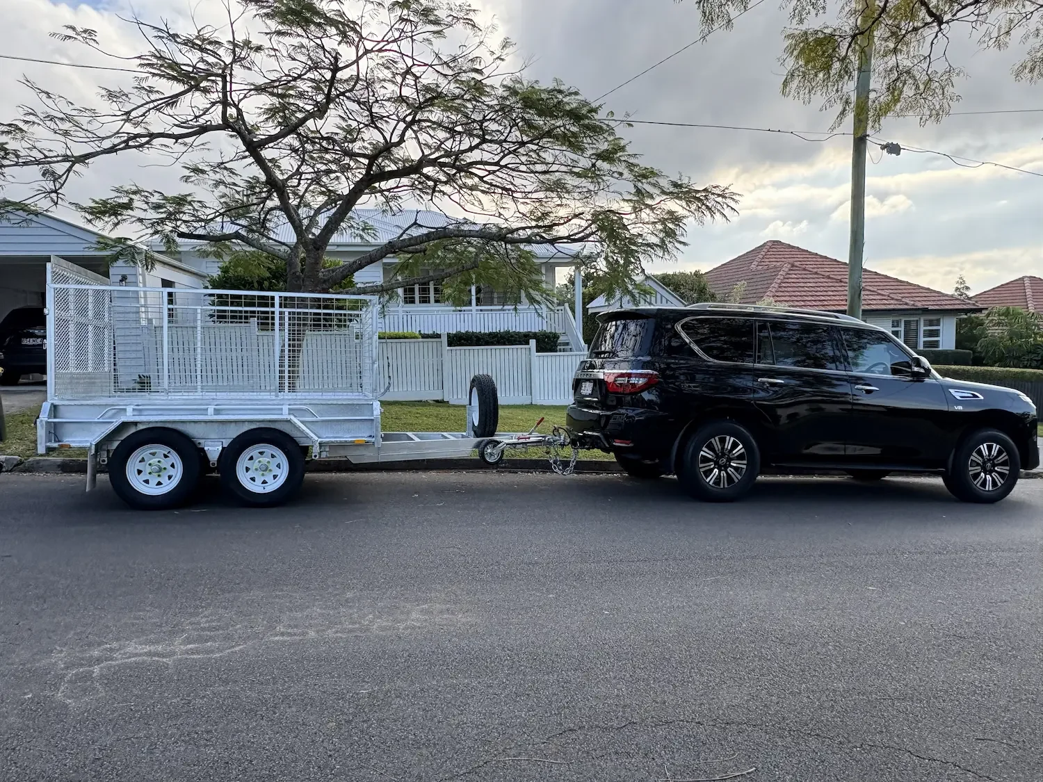 A black SUV parked on the street attached to an aluminum trailer with a cage, a spare tire, and chains, with a tree and residential houses in the background.