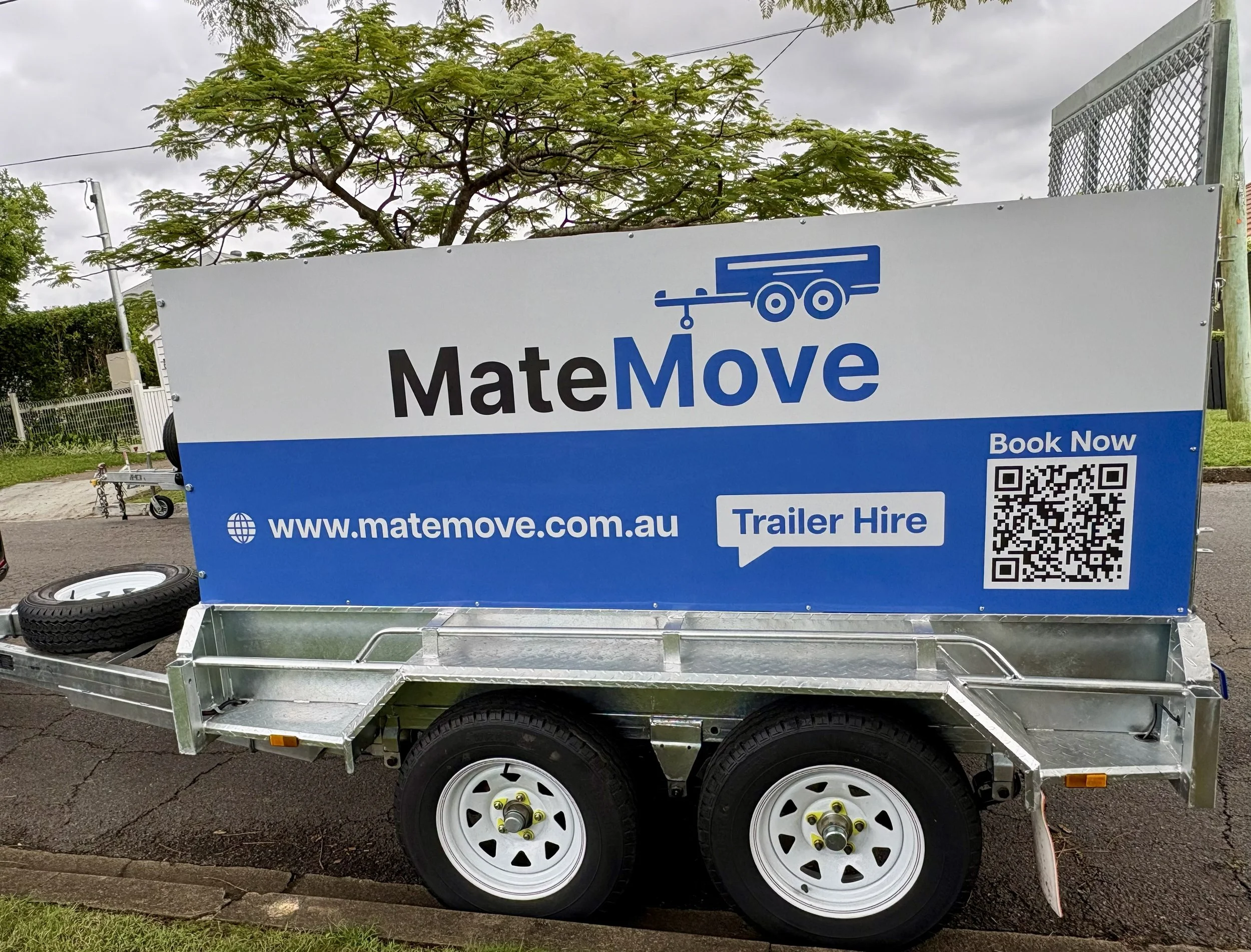 A trailer with the MateMove logo and website, parked on a street with trees and cloudy sky in the background. The trailer has a QR code and text indicating trailer hire and booking options.