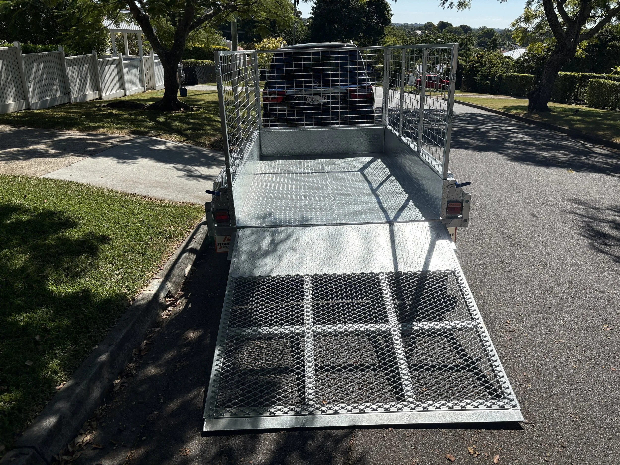 A metal flatbed trailer parked on a street with trees and a white fence in the background.