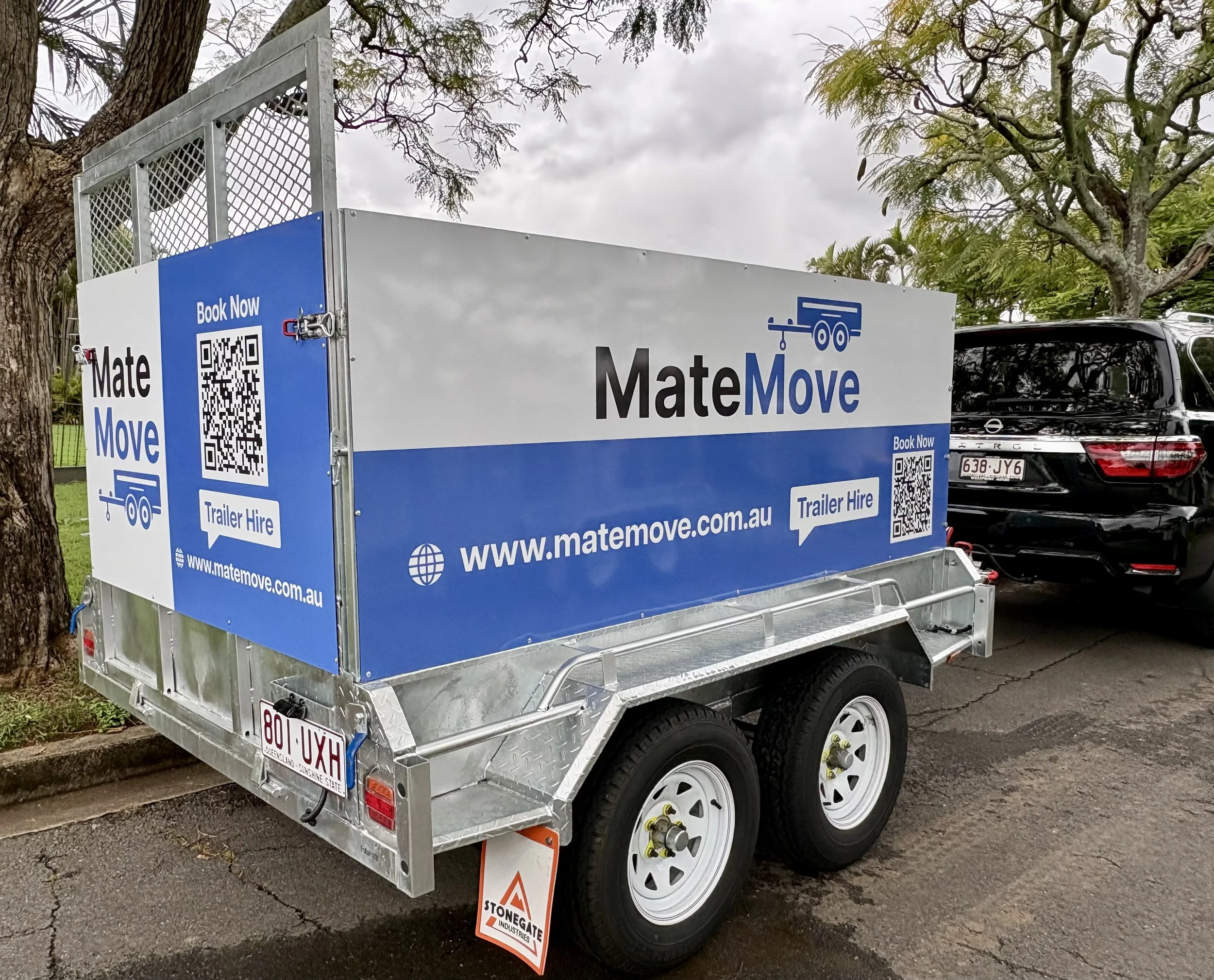 A small silver trailer with advertising for MateMove shows a QR code and website URL. It is parked on the street under tree branches with overcast sky. The trailer has a sign indicating trailer hire services and the company logo.