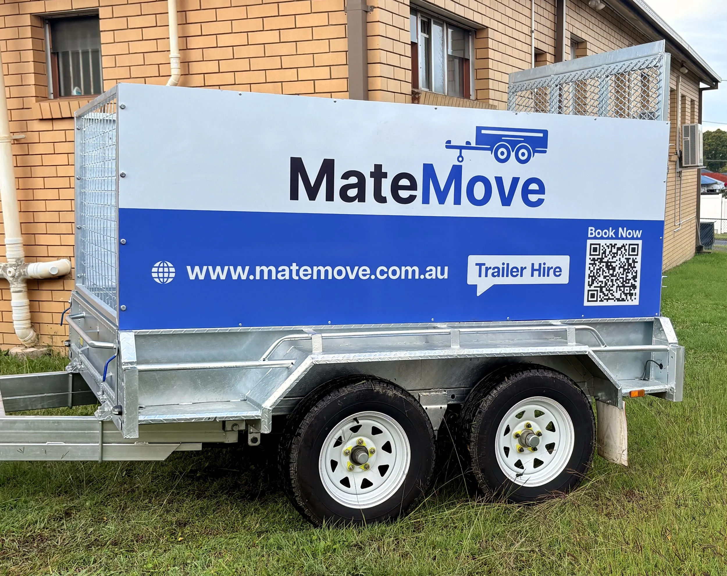 A silver utility trailer parked on grass in front of a brick house, displaying a large white and blue advertisement sign for MateMove, a moving and trailer hire company, with a QR code and website URL.