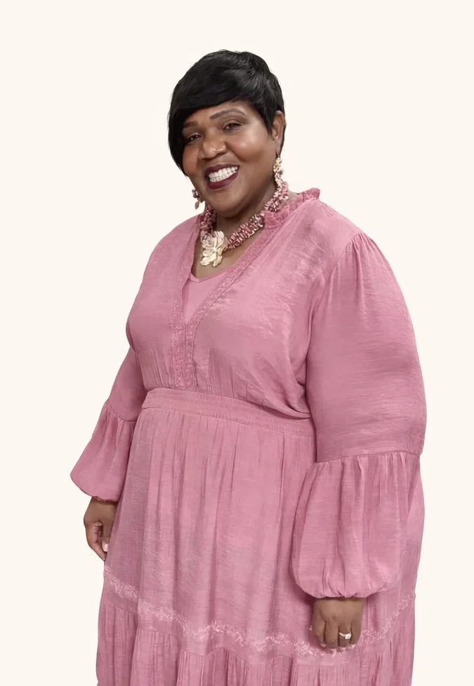 Portrait of a woman with short black hair wearing a pink dress with puffed sleeves, accessorized with jewelry, standing against a plain background.