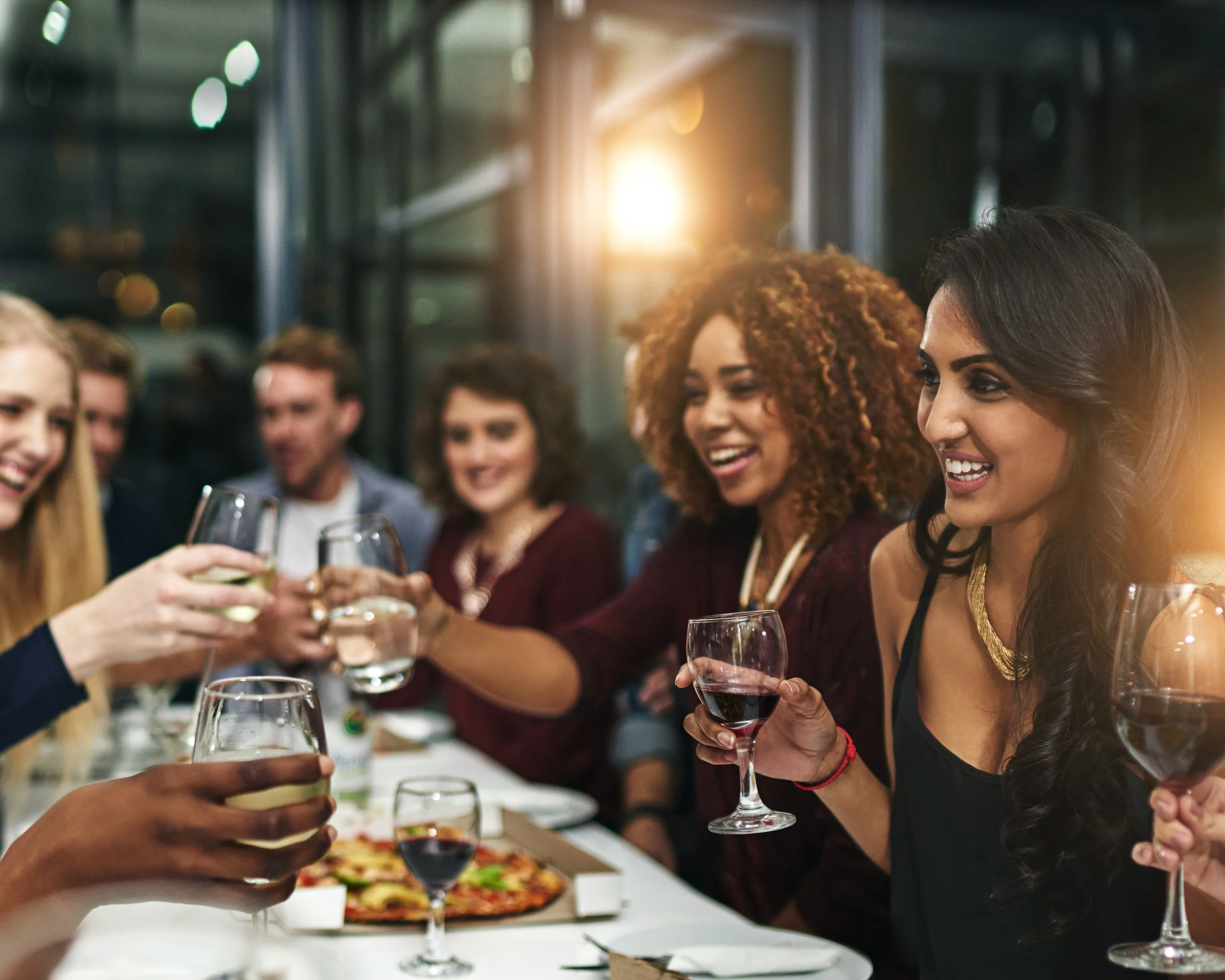 A group of friends enjoying drinks and toasts at a dinner table, with a pizza in the center.