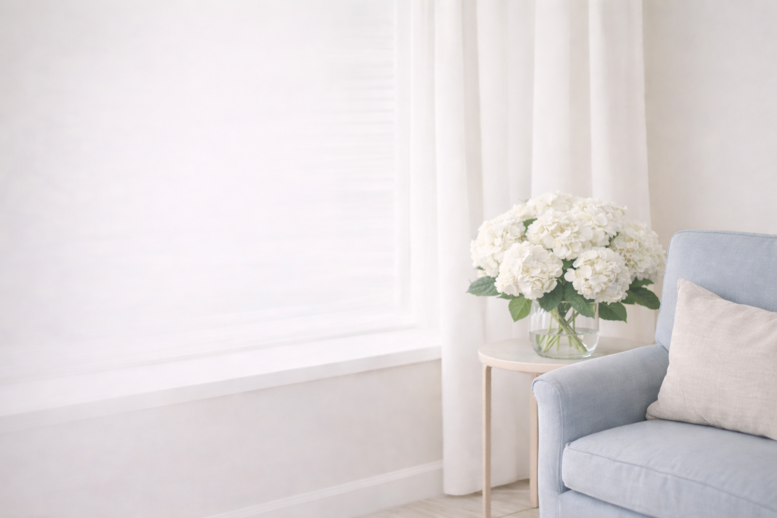 A living room corner with a light blue armchair, a white pillow, a small round white table with a glass vase of white hydrangeas, and white curtains and window blinds in the background.