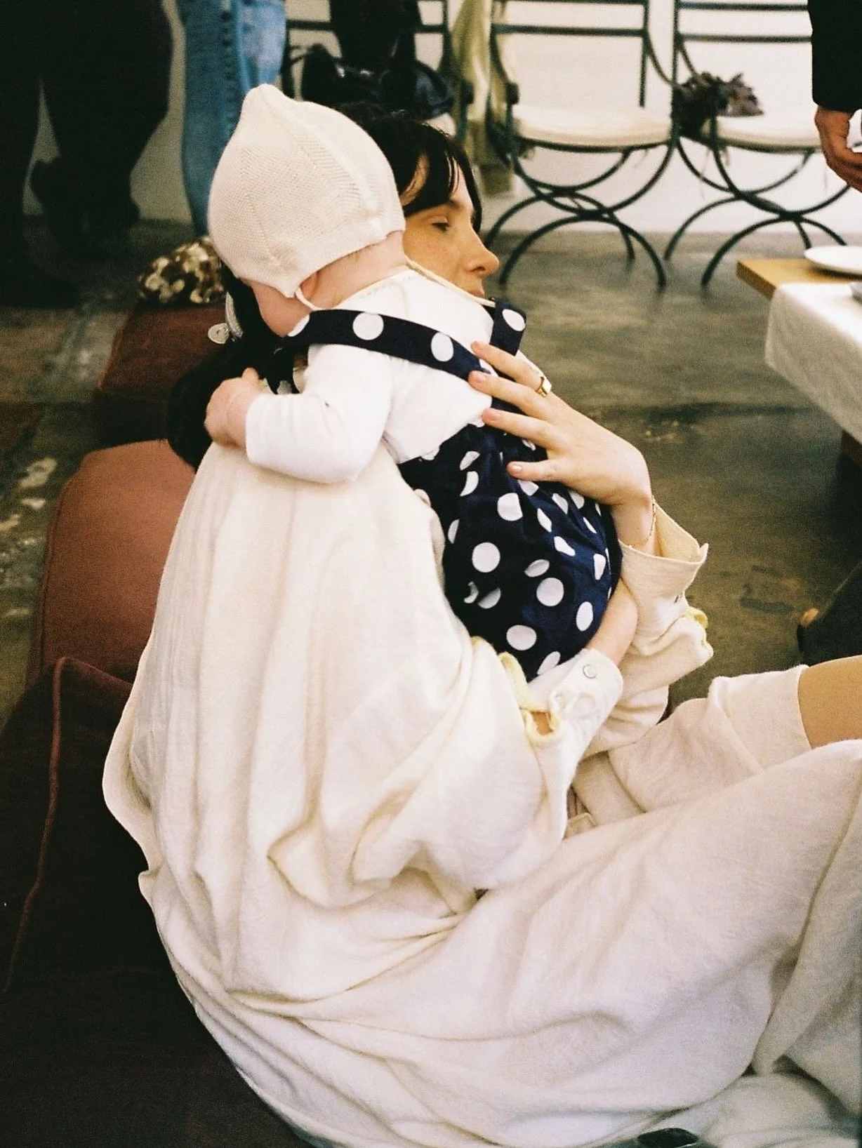 A woman sitting on the floor with a small child hugging her neck. The woman has dark hair and is wearing a light-colored long-sleeve shirt, while the child is wearing a white hat, a white outfit, and a navy blue polka-dot vest. The woman's eyes are closed, and she appears to be relaxed and content.