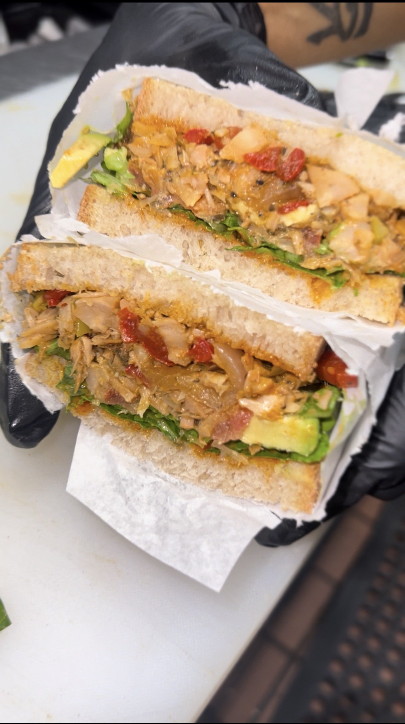 Two halves of RHW Jerk Chkn jackfruit sandwich with peppers and greens held in a black-gloved hand