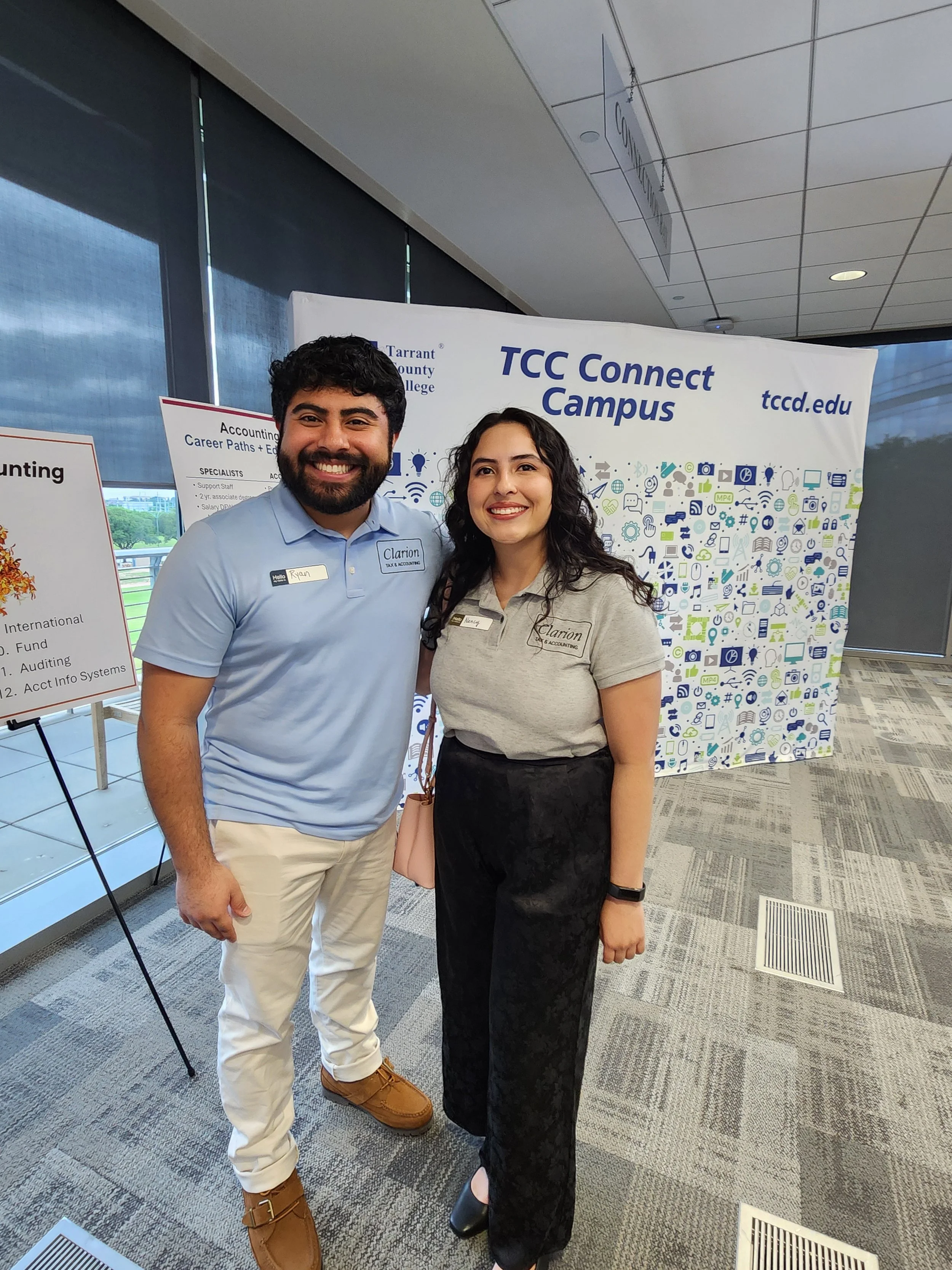 Two young adults, a man and a woman, smiling and standing close together in front of a TCC Connect Campus display, in an indoor setting with large windows and modern interior design.