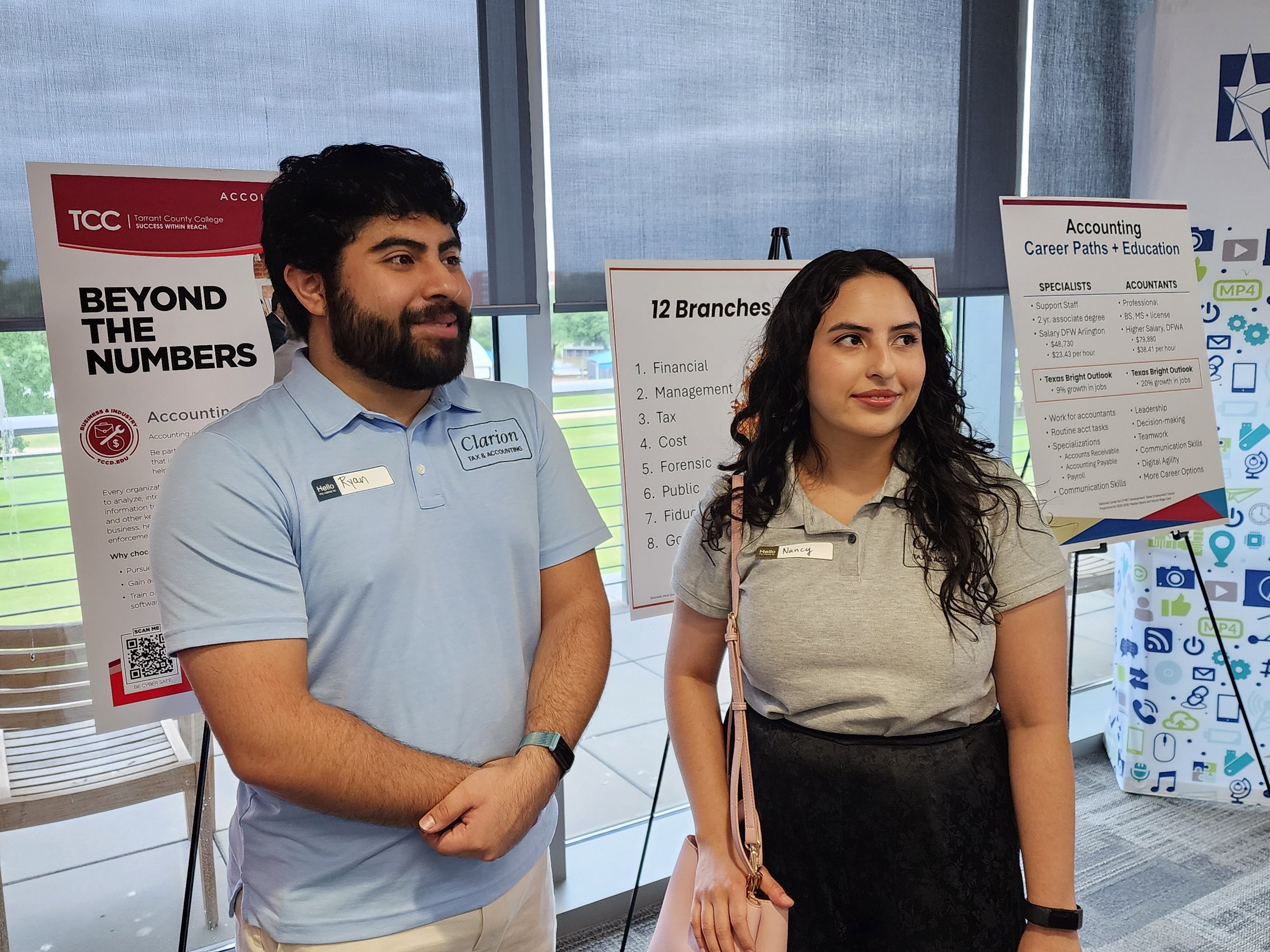 Two young adults, a man with a beard and a woman with curly hair, standing in front of informational posters at an event. The man wears a light blue shirt and has a name tag reading 'Ryan'. The woman wears a beige shirt and has a name tag reading 'Nancy'. Behind them are posters about accounting career paths and a college.