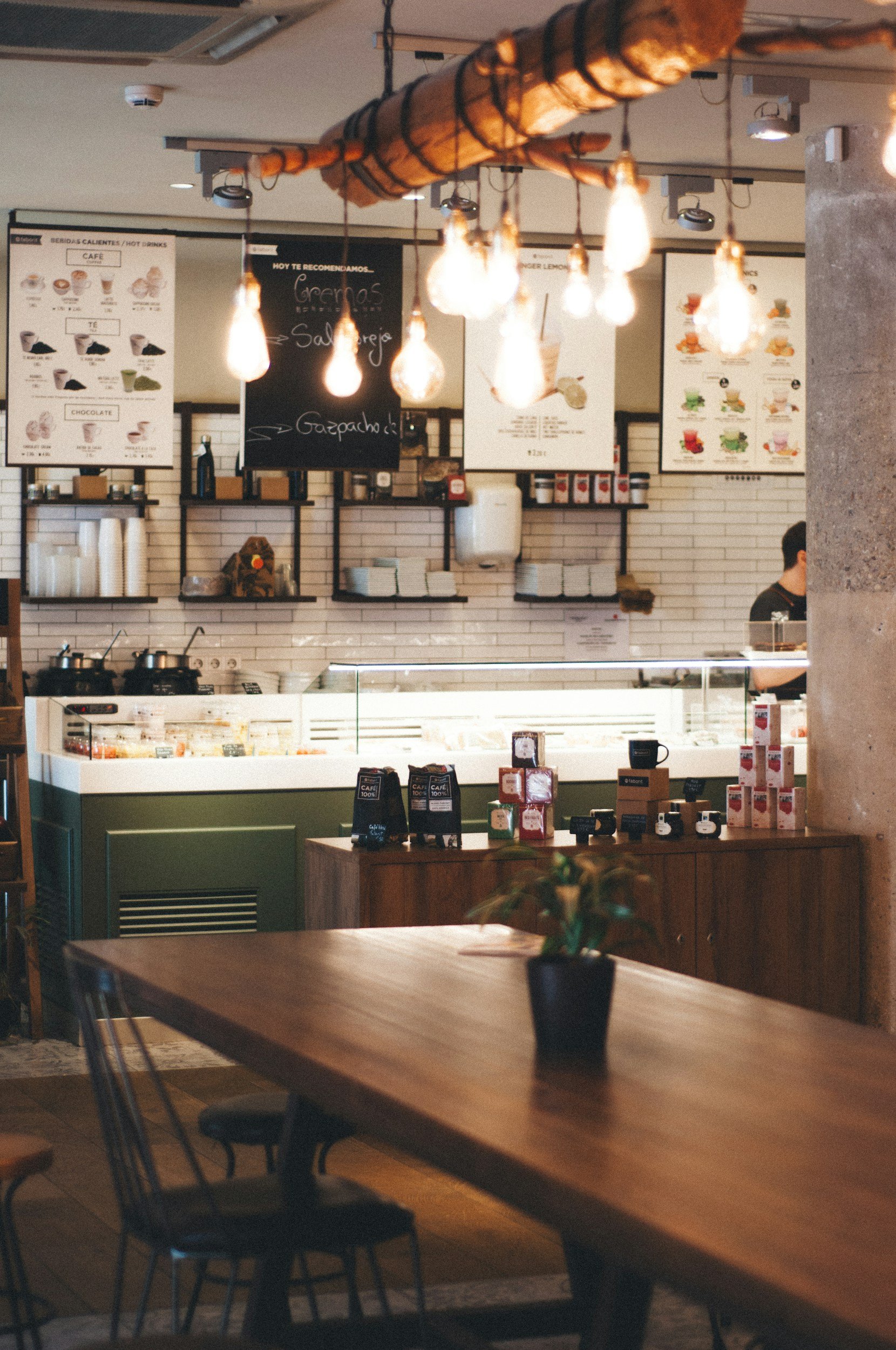 Interior of a coffee shop with a wooden table and chairs in the foreground, a counter with various items, and menu boards hanging on the wall in the background. Hanging light bulbs are also visible.