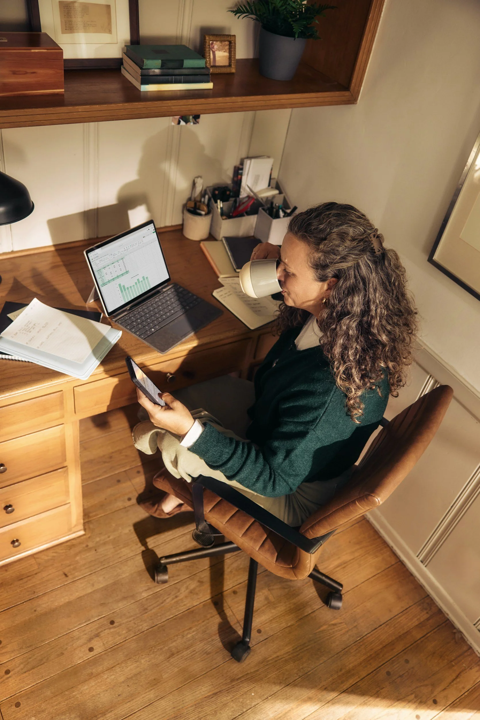 A woman with curly hair, wearing a green jacket, sitting in a brown office chair with a black armrest in a home office, holding a smartphone. She has a coffee mug in her face, a laptop with a spreadsheet and chart visible on the screen, a notebook, and a stack of papers in front of her. The desk has a wooden surface, with a built-in drawer underneath. Behind her, there are shelves with books, plants, boxes, and folders. The room has wooden flooring and white walls with framed pictures.