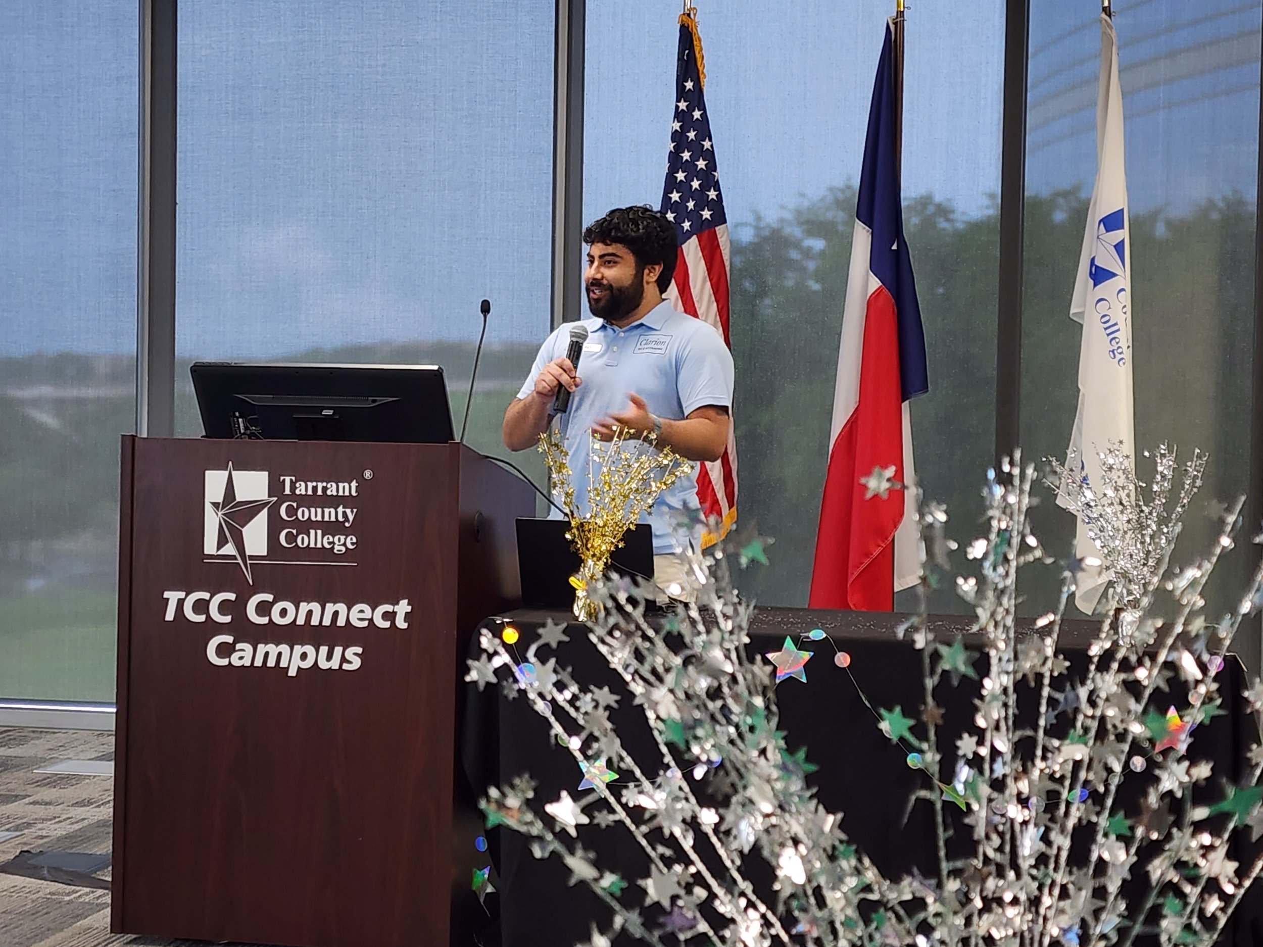 A young man speaking at a podium with a microphone, in front of flags representing the United States and Texas, at Tarrant County College, TCC Connect Campus.