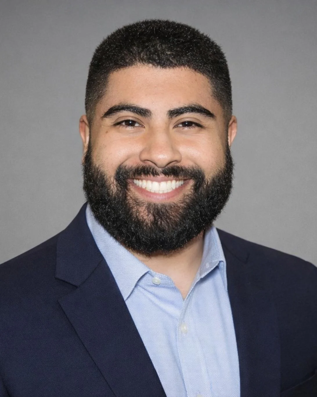 Headshot of a smiling man with a dark beard and short dark hair, wearing a navy blazer and light blue shirt against a gray background.
