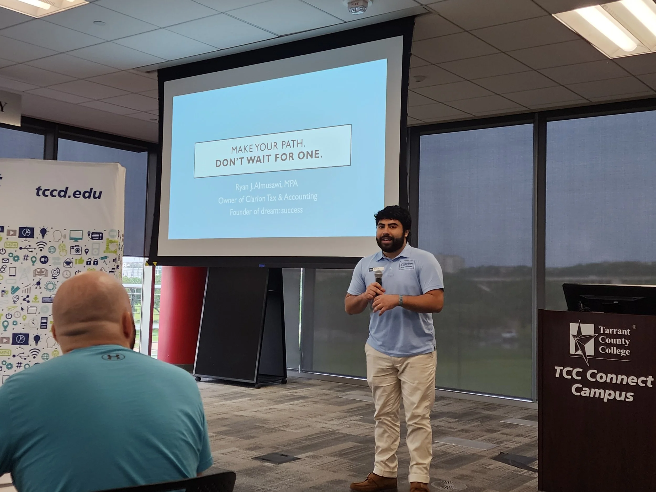 A man with dark hair and a beard stands holding a microphone in front of a large screen with a presentation slide that reads, "Make your path. Don’t wait for one." The slide also displays the presenter's name, Ryan J. Almusawi, MPA, along with his credentials and affiliations. The background shows a conference room with windows, and there are several attendees present, including a person in a turquoise shirt with their back to the camera.