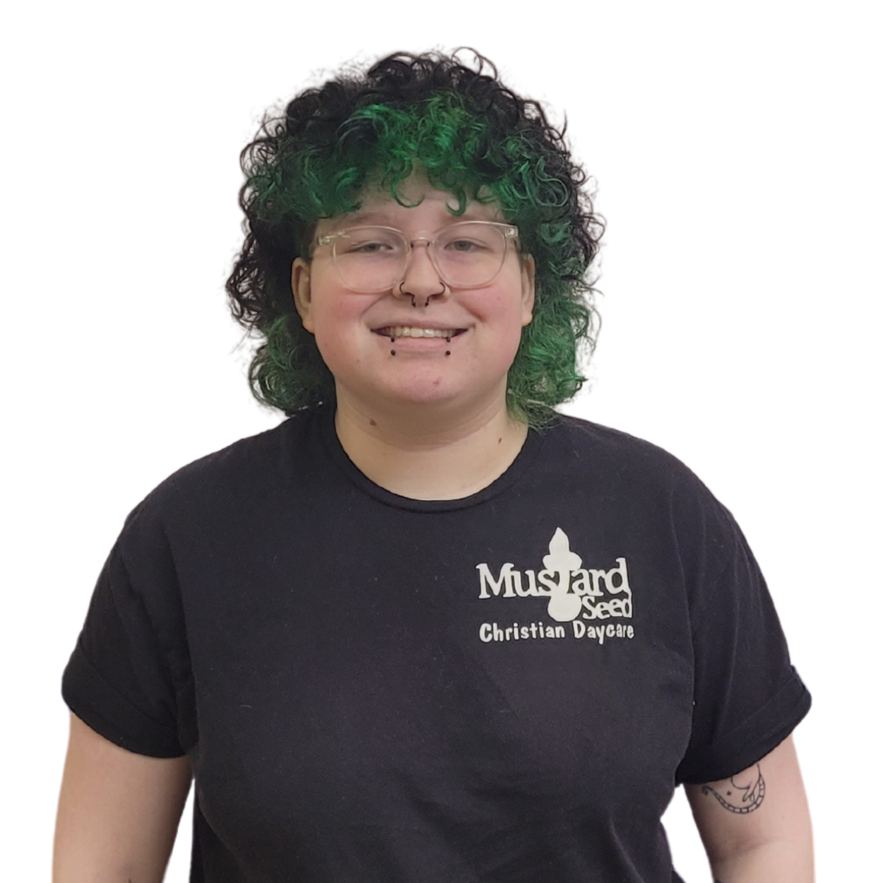 Person with curly black and green hair wearing glasses and a black t-shirt with a mustard seed logo and Christian daycare text, smiling, against a white background.