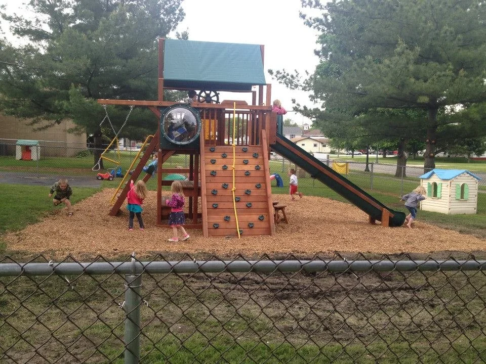 Children playing on a wooden playground with a slide, climbing wall, and swings in a park surrounded by trees and a chain-link fence.