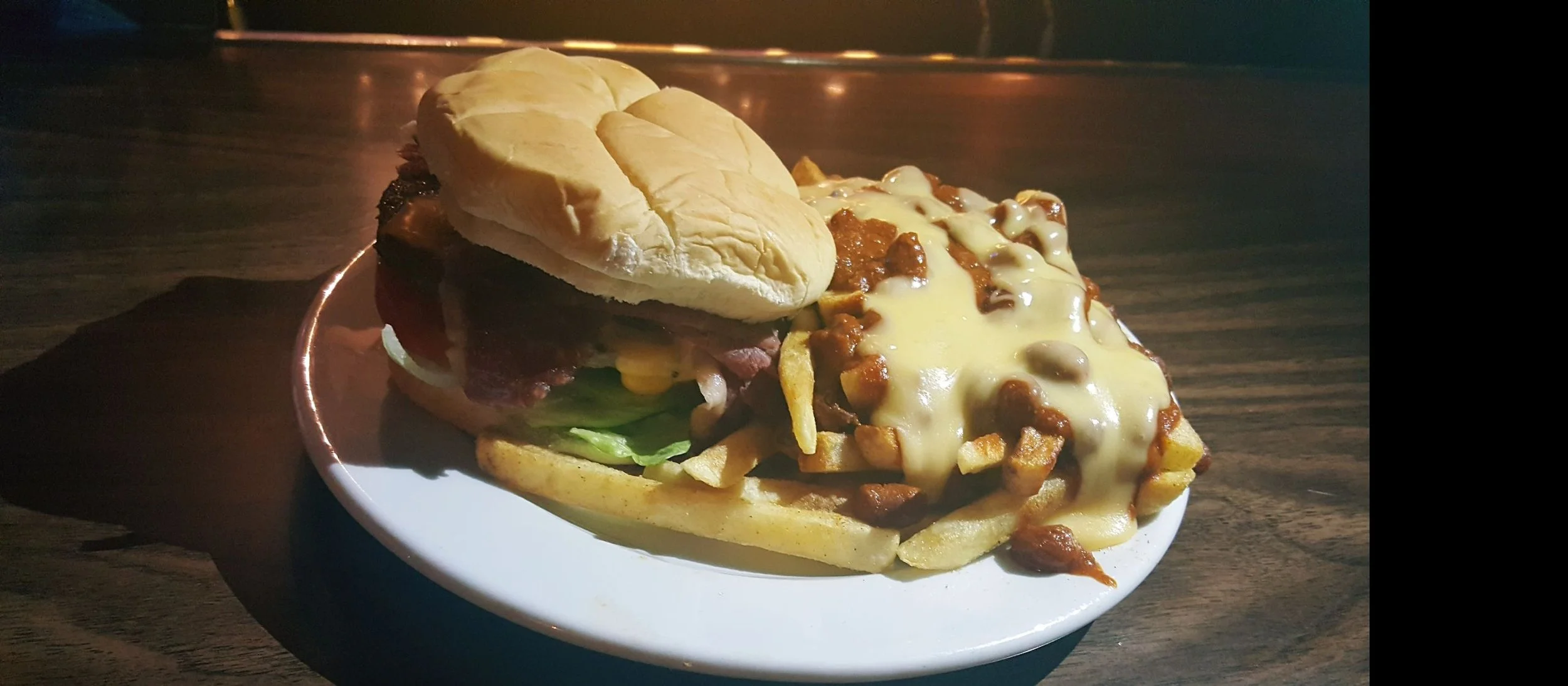 A plate of a cheeseburger with lettuce, tomato, and onions on a bun, served with a side of loaded French fries topped with cheese and chili.
