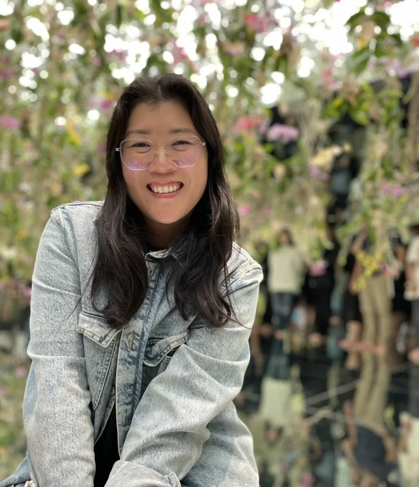 A woman with long dark hair, glasses, and a light denim jacket, smiling and sitting in front of a flowering tree with pink and white blossoms, with people in the background.
