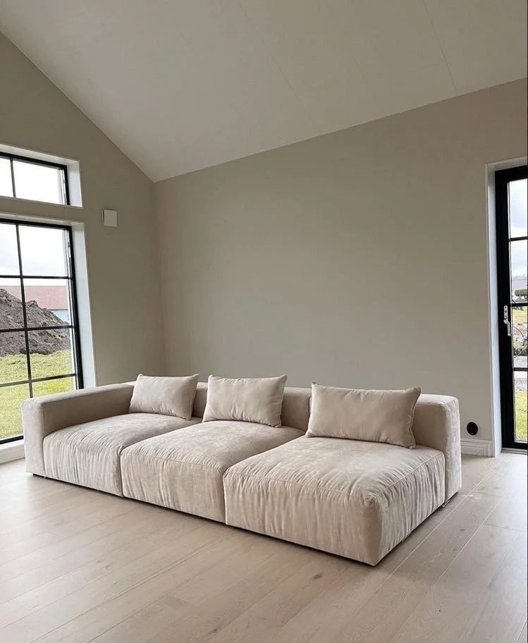 A beige sofa with three matching pillows in a modern, minimally decorated living room with large windows and light-colored flooring.