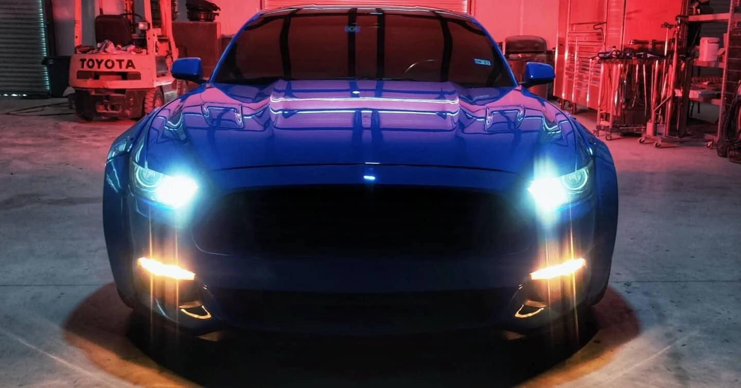 Black sports car with headlights on, parked inside a garage with tools and equipment, illuminated by red and white lights.