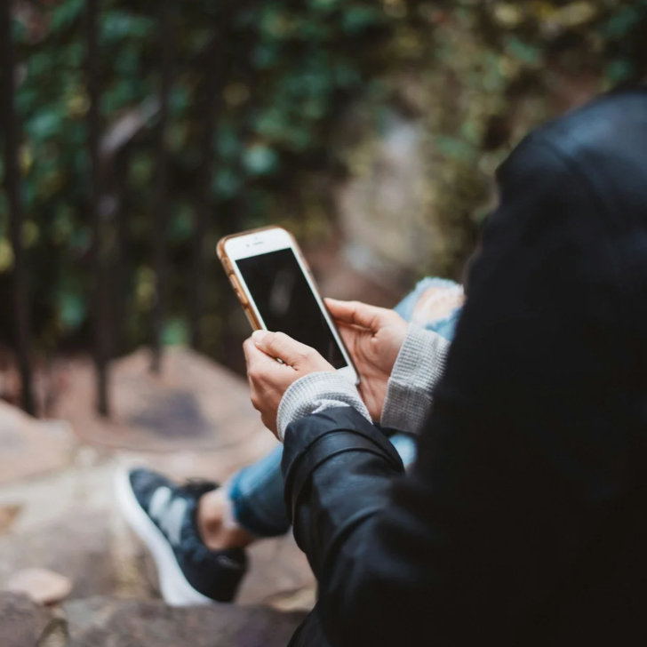 Person sitting outdoors on a rocky surface, using a smartphone, with a blurred natural background.