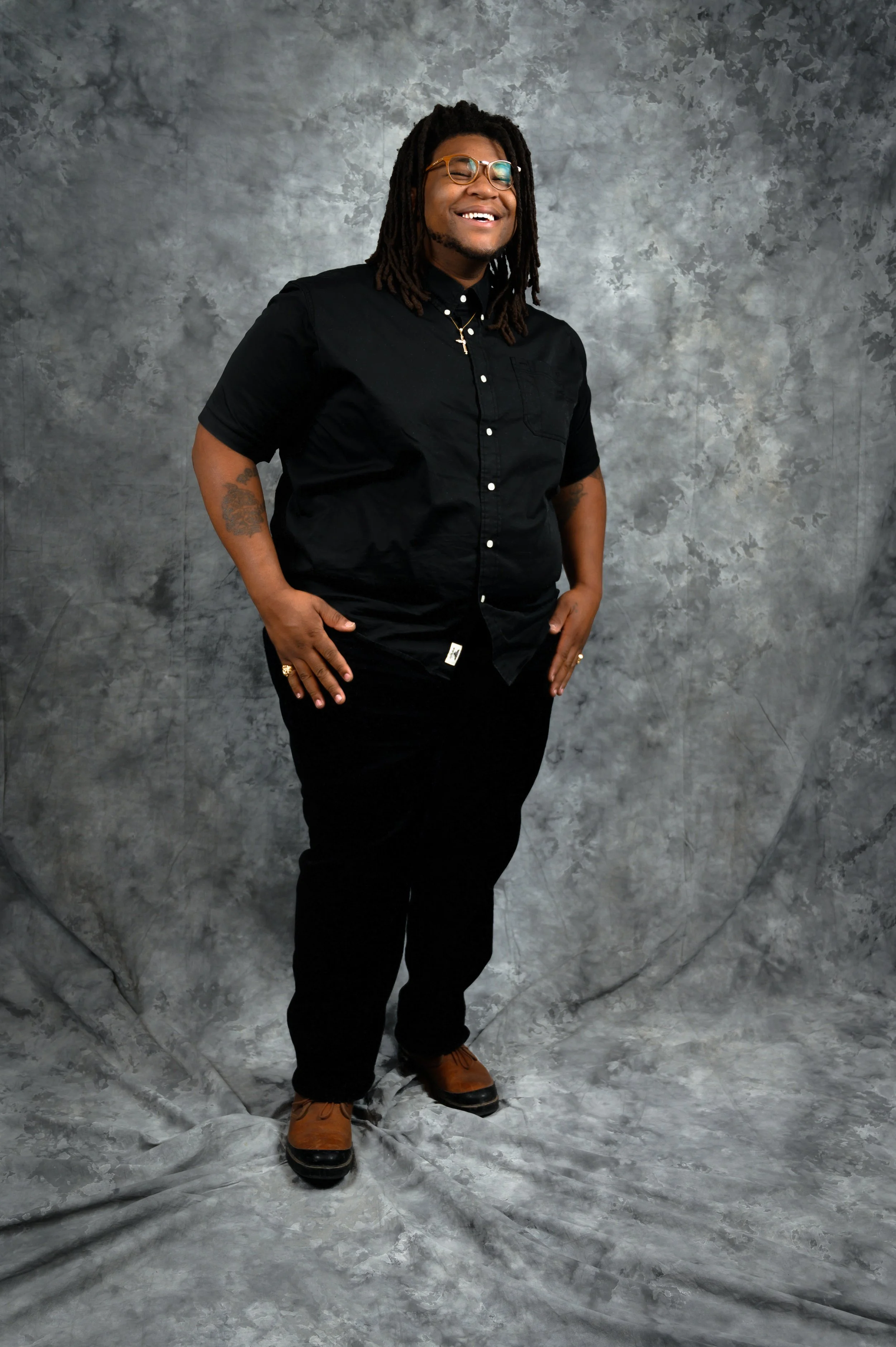 A man Deacon Phil or Philip Israel with glasses, dreadlocks, and tattoos, dressed in a black shirt and black pants, smiling and standing with hands in pockets against a gray textured backdrop.