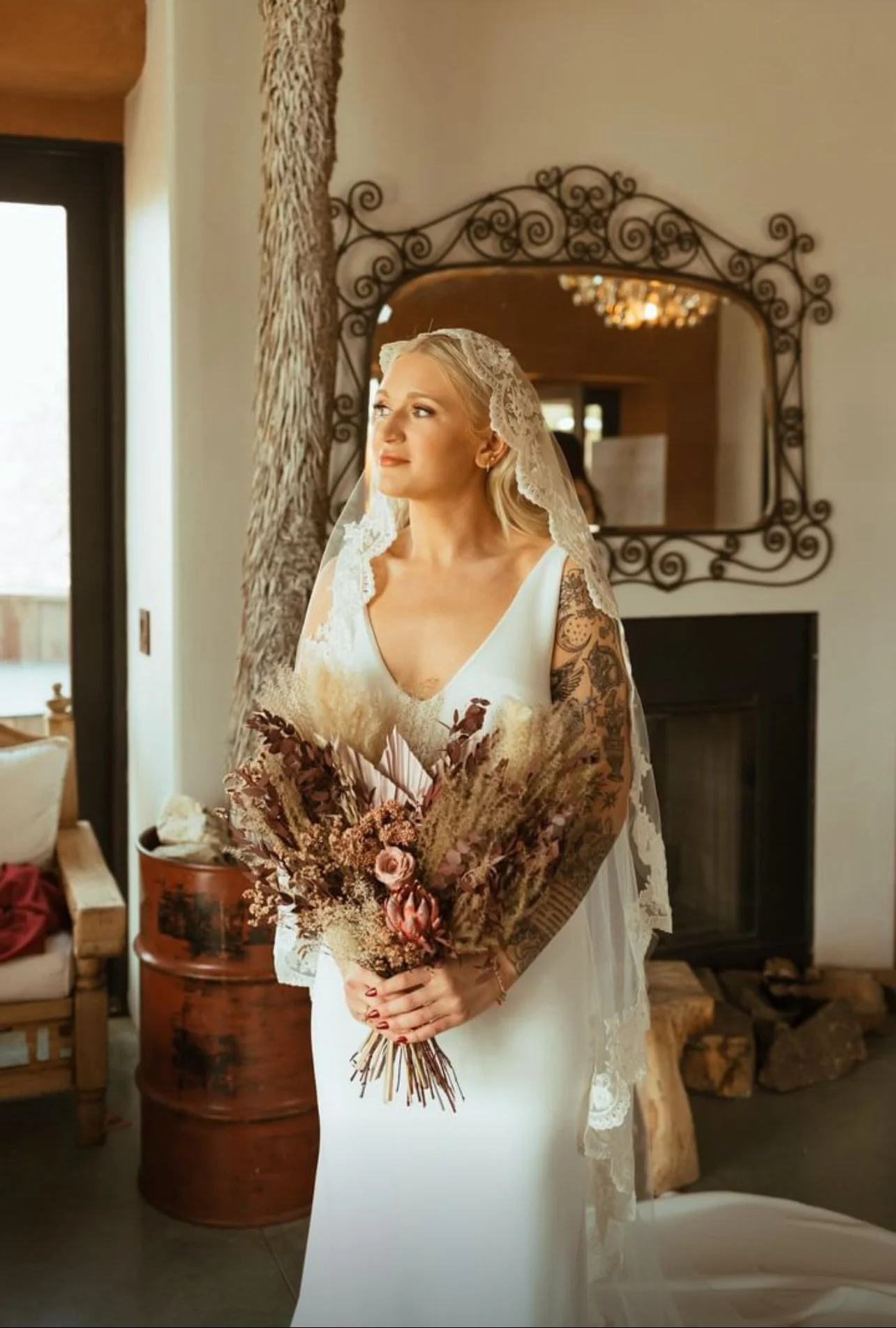 A bride in a white wedding dress holding a bouquet of dried flowers, standing indoors near a fireplace with a mirror above it, decorative wrought iron. She wears a lace veil and has tattoos on her arms, looking thoughtfully to the side.
