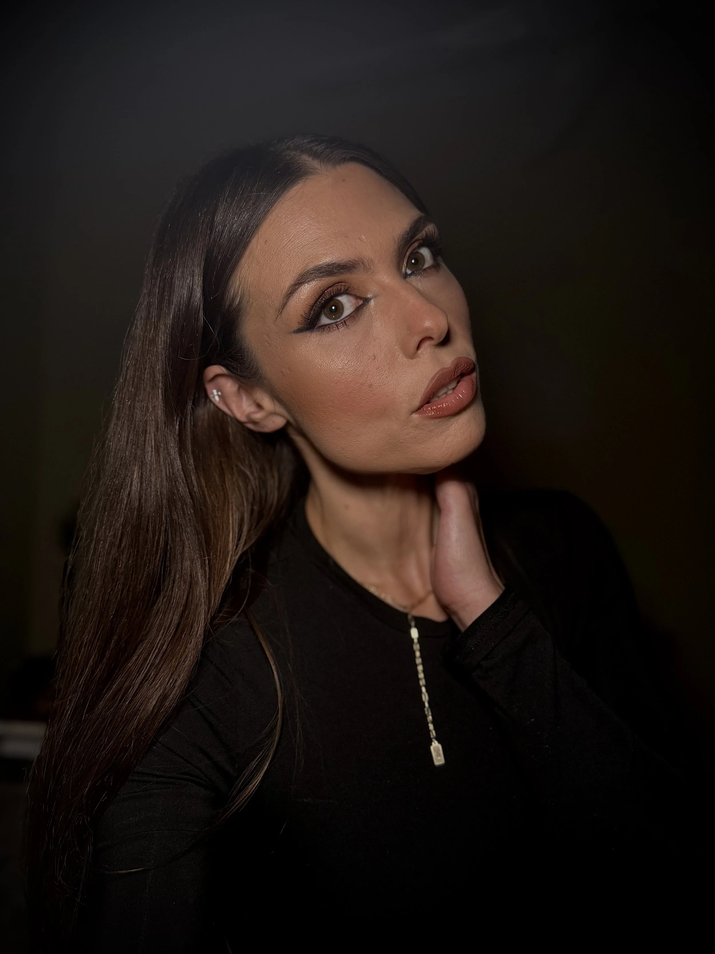 Portrait of a woman with long brown hair, wearing makeup, a black top, and a gold necklace, posing with her hand on her neck against a dark background.