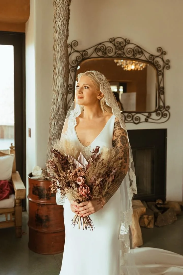 A bride with blonde hair in a white wedding dress and lace veil holding a bouquet of dried flowers, standing in a warmly lit room with a decorative mirror on the wall.