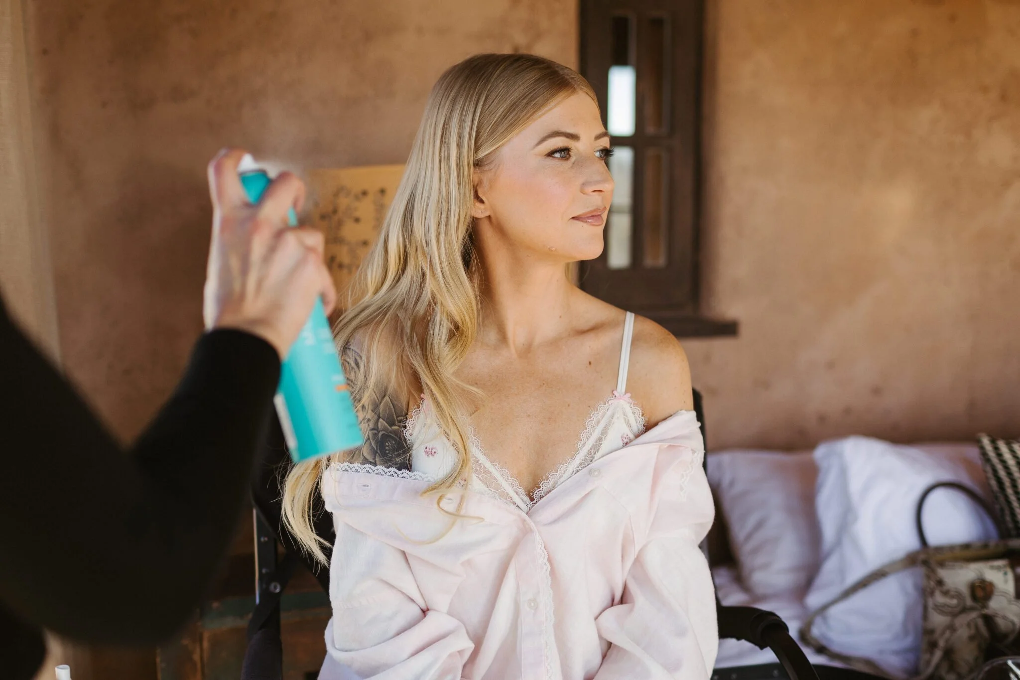 A woman with long blonde hair sitting in a room, looking to the side, while someone sprays her face with a spray bottle.