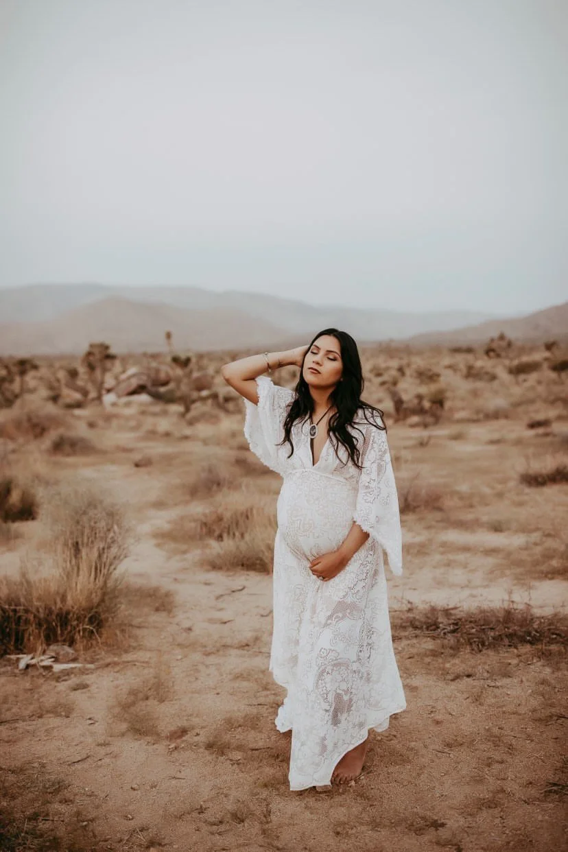 Woman in hair and makeup by Danielle Rose Beauty Atelier. Her hair is styled in loose effortless waves, and her makeup is soft. She is wearing a lace bohemian white gown paired with a bolo necklace. She is grasping her pregnant belly looking peaceful