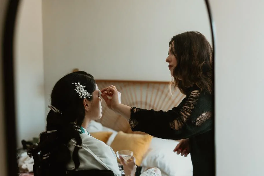 A makeup artist applying makeup on a woman sitting on a bed.