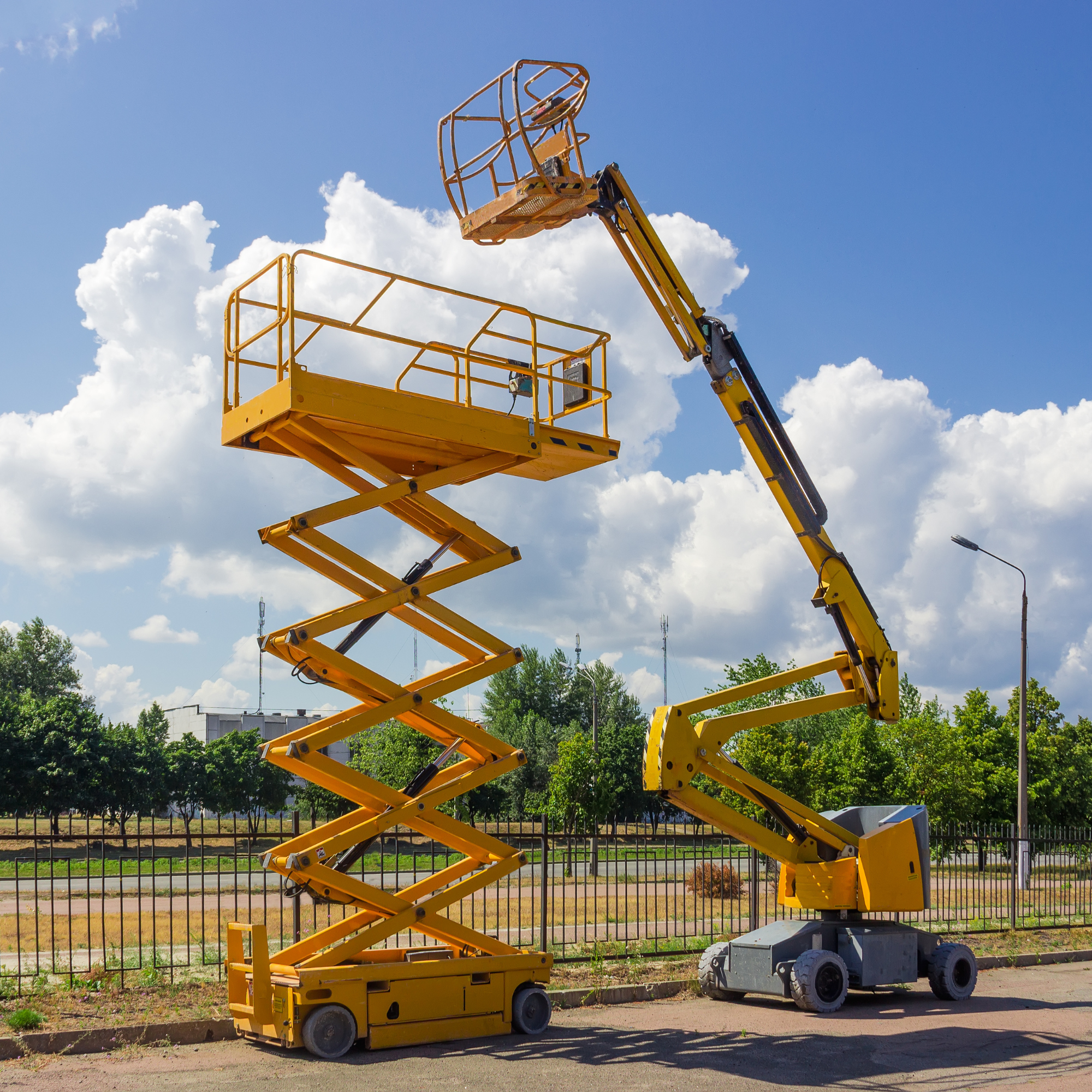 A yellow and gray telescopic boom lift with a yellow work platform raising into the sky, with trees and a fence in the background under a partly cloudy sky.