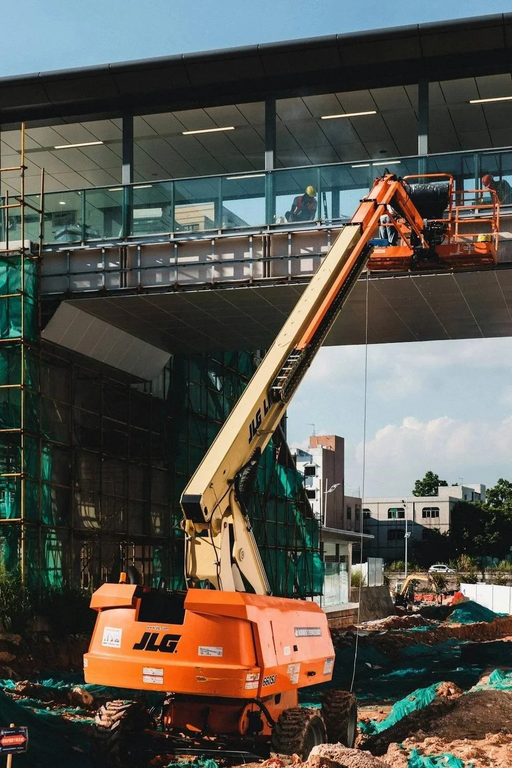 A construction site with an orange JLG lift extending to a balcony of a building. Workers with safety helmets are on the balcony, working on the railing. The building has glass windows and an overhang. Green safety netting surrounds parts of the cons