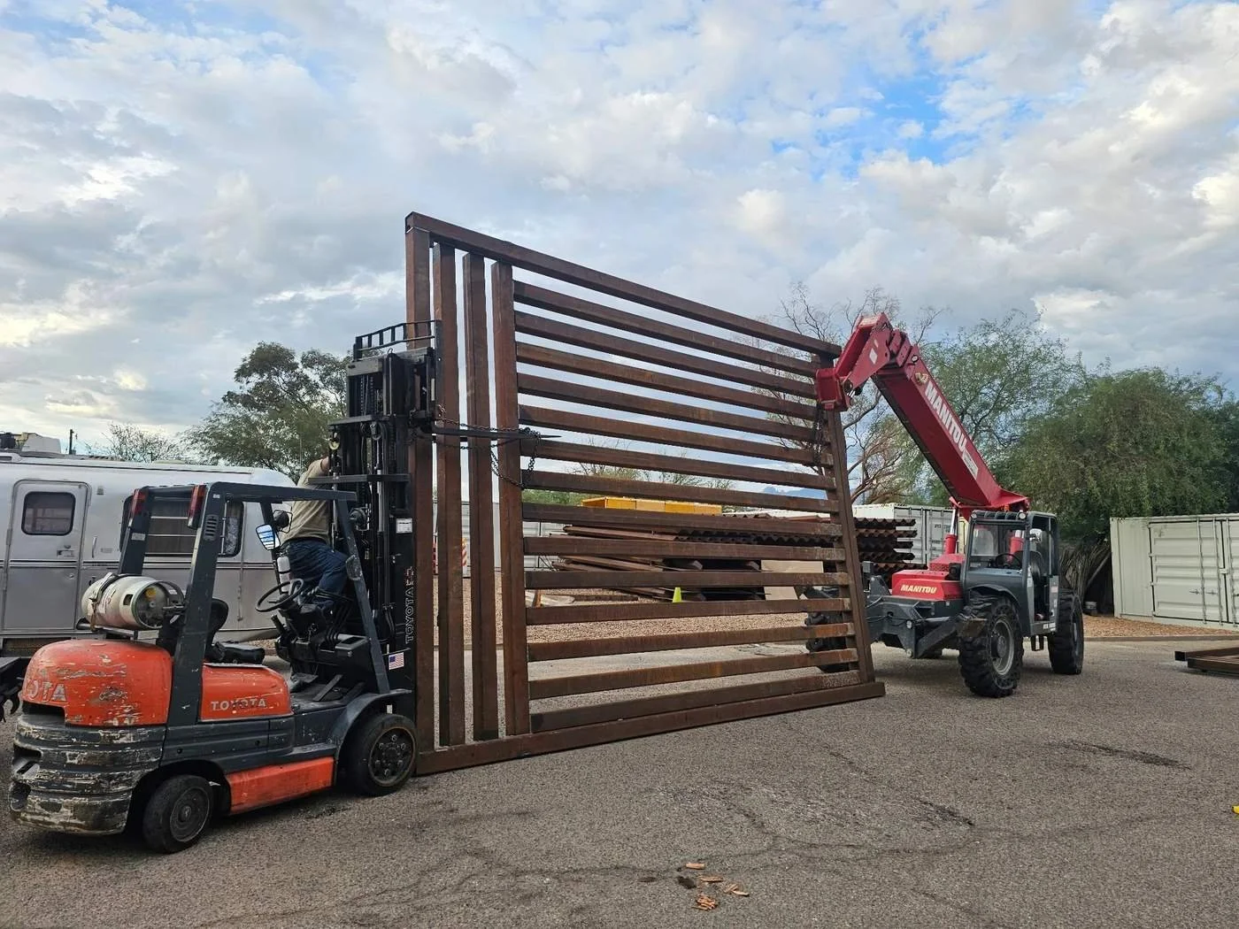 A forklift and a crane are lifting and moving a large wooden gate in an outdoor lot with trees, a trailer, and cloudy sky in the background.
