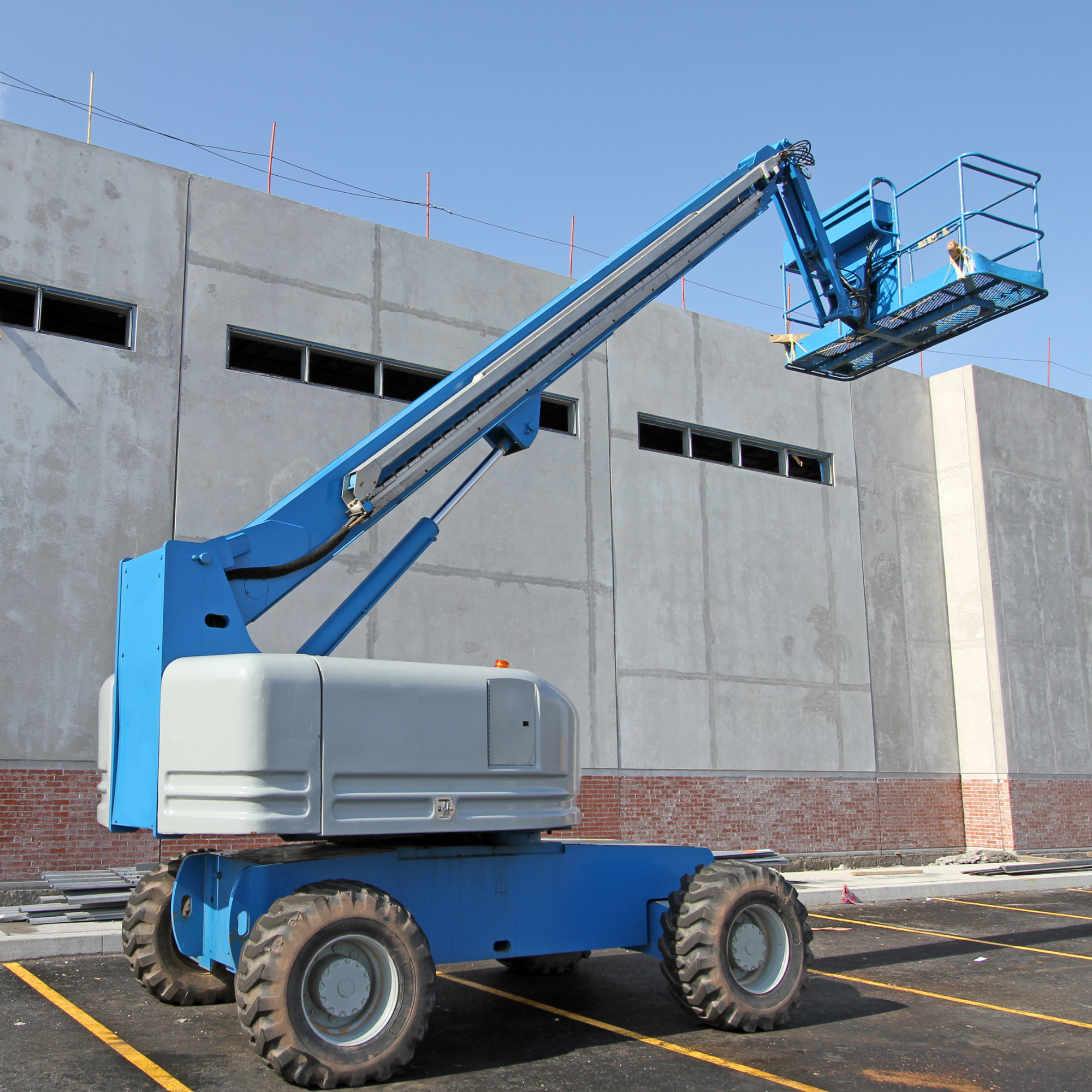 A blue mobile elevating work platform (MEWP) with a basket, parked in a paved parking lot near a building under construction or renovation, with a clear blue sky above.