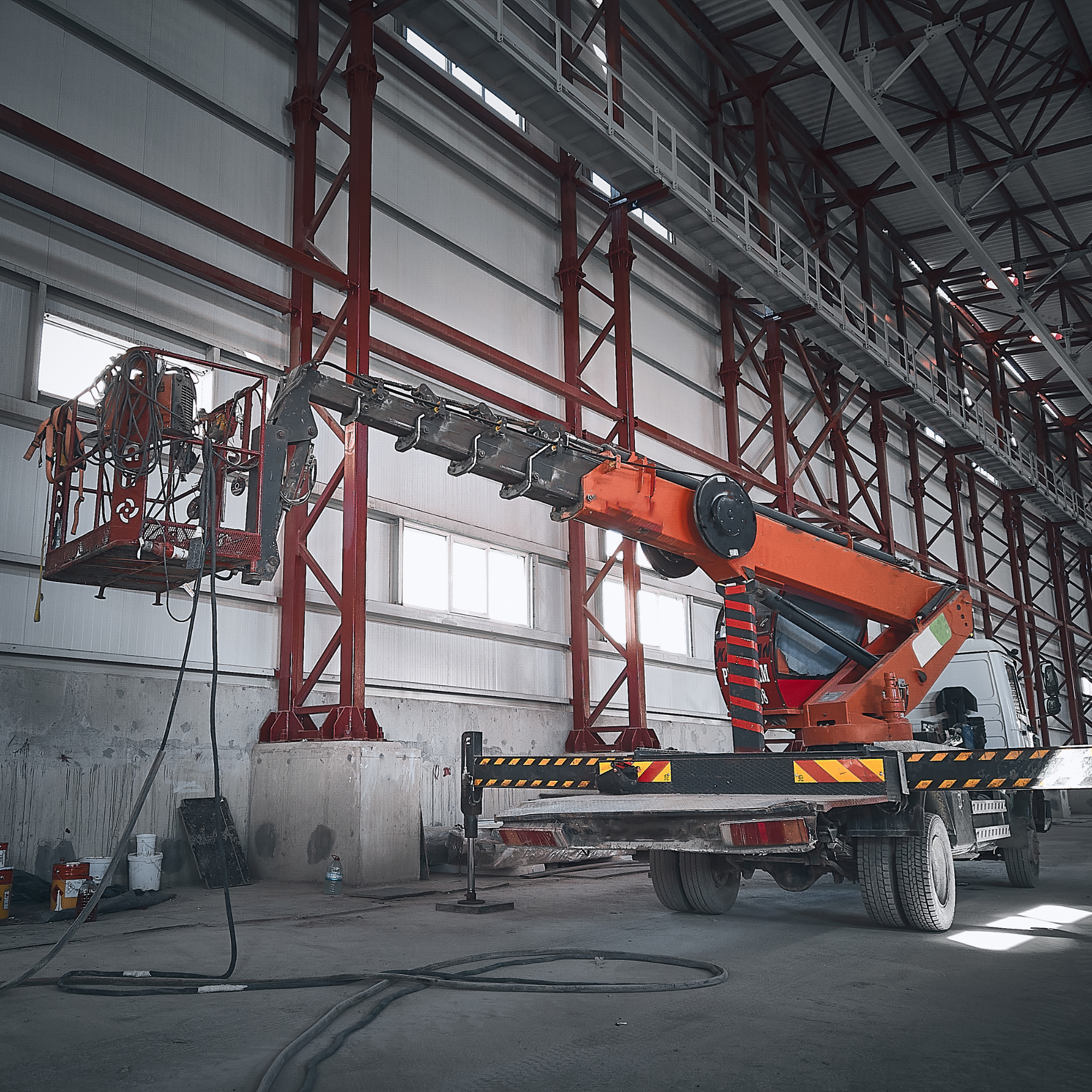 Construction site with a cherry picker lift and large orange crane inside a warehouse, with visible scaffolding, tools, and construction materials scattered on the floor.