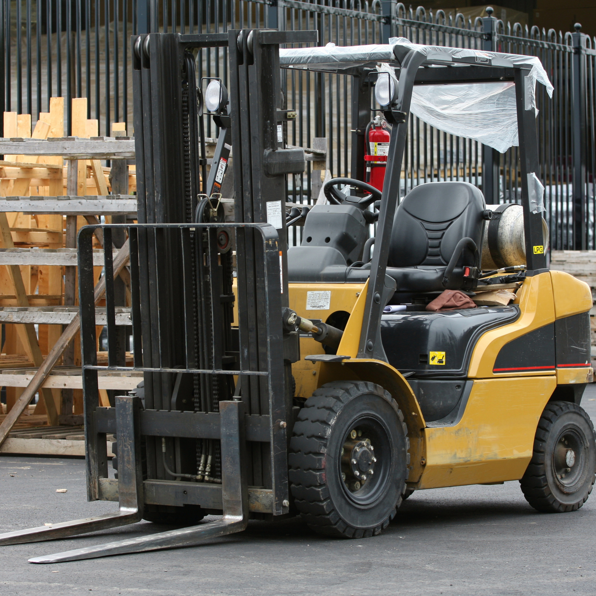 A yellow and black forklift parked outdoors, with a black safety cage, on a paved surface, surrounded by a black metal fence and wooden pallets stacked in the background.